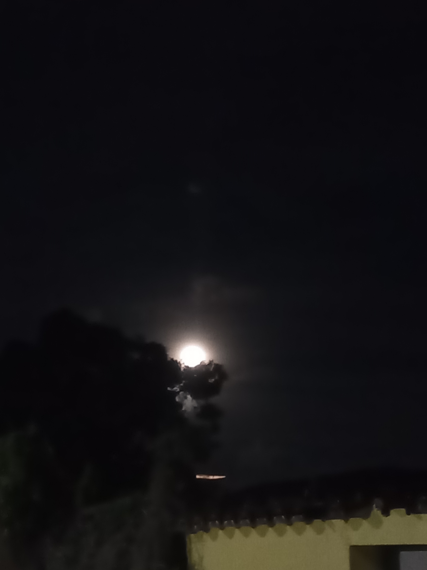 At night, the moon is visible through a scattering of clouds over Juan Griego, Venezuela. A dark tree stands to the left of the moon, while a light-colored building with a ridged roof is visible in the lower right corner of the frame. The sky is otherwise dark and clear.
