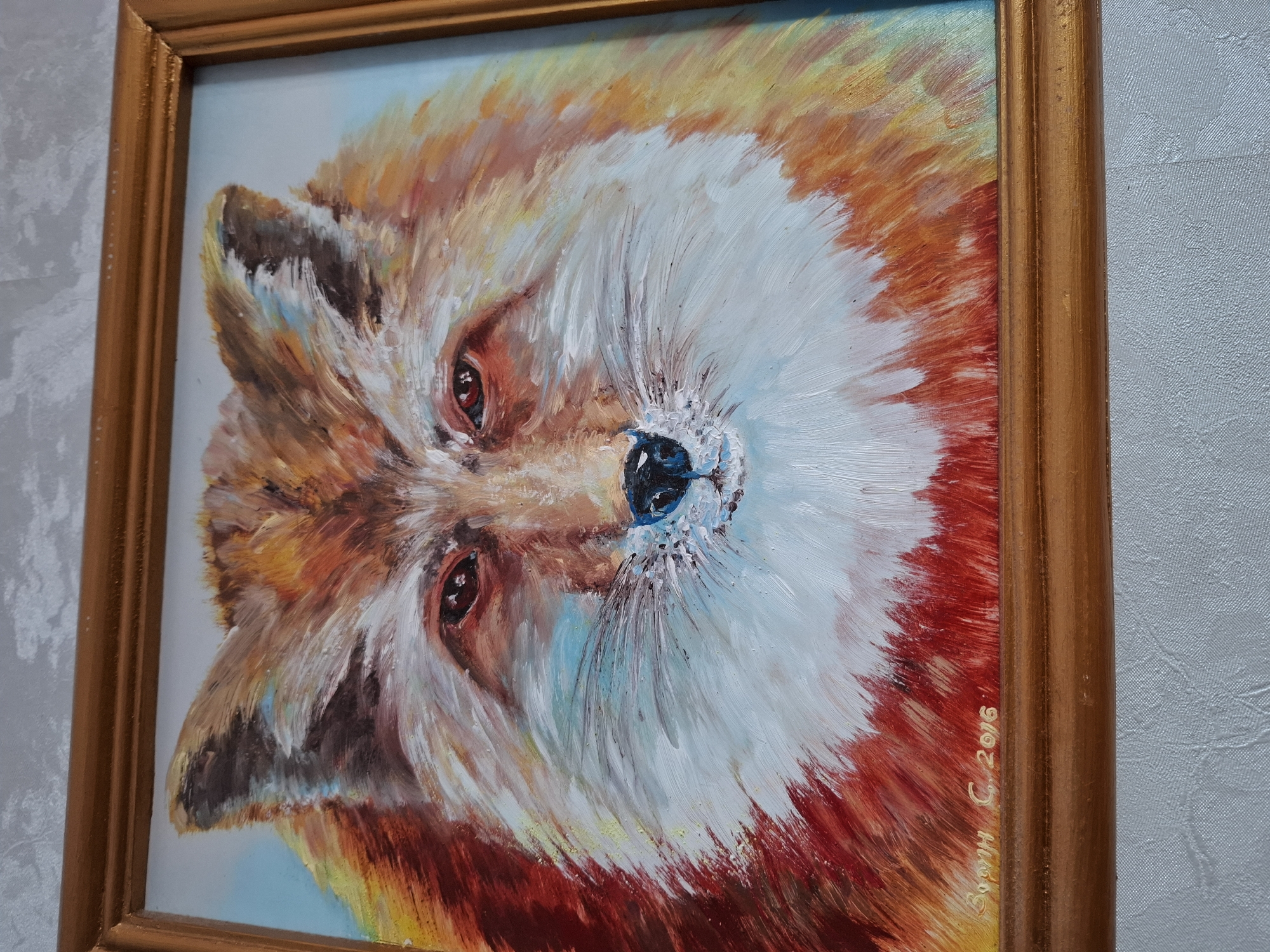 The image depicts a framed oil painting of a fox head. The fox has a predominantly white face with orange and brown fur around the perimeter. The background is light blue. The painting has a gold-colored frame. The painting appears to be photographed indoors against a light gray textured wall. "Вульф С. 2016" is signed on the bottom right of the painting. The location is Chaikovsky, Russia.
