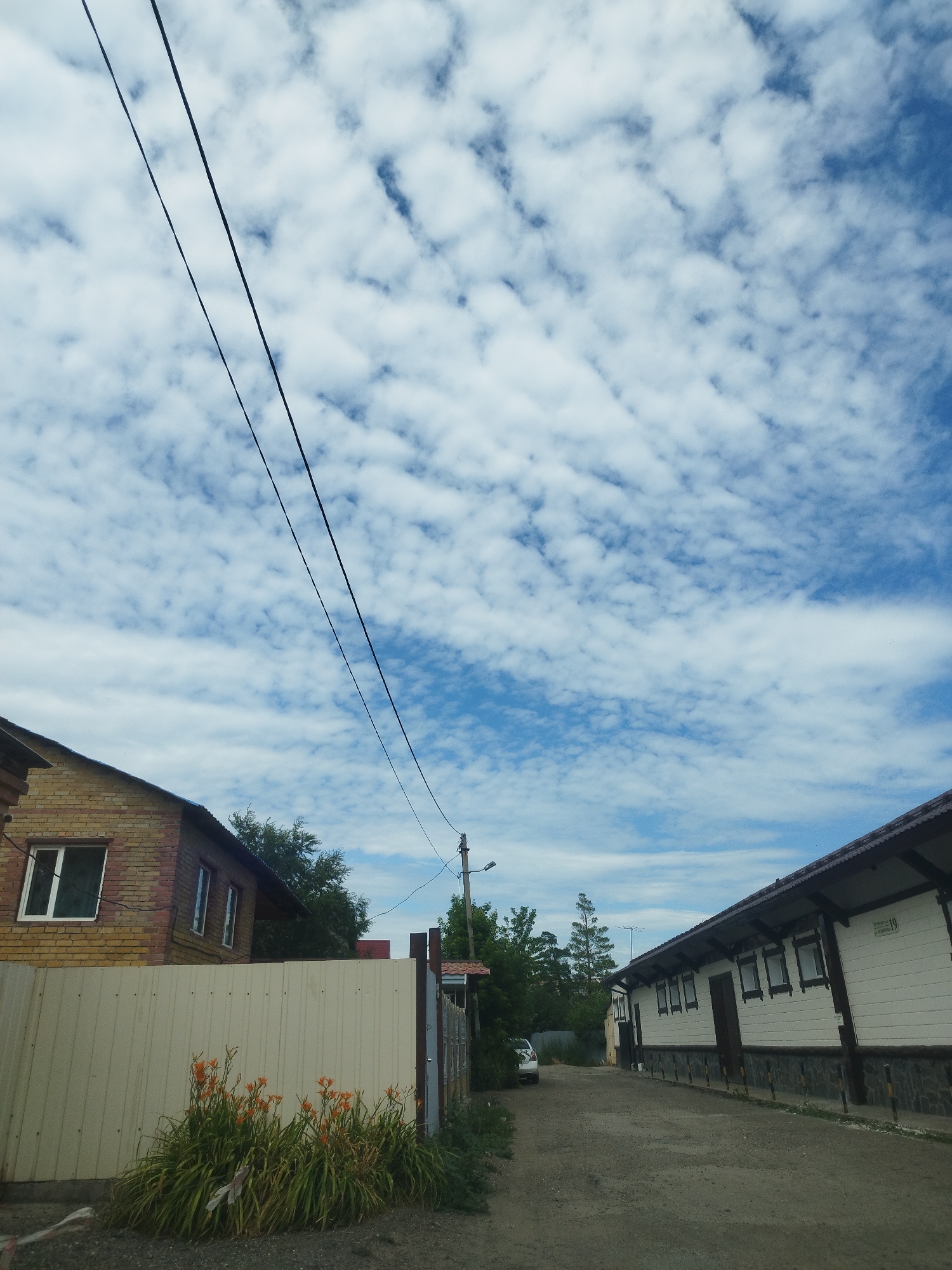 The image depicts a street scene in Oskemen, Kazakhstan. On the left, a two-story brick house with white-trimmed windows is visible behind a light-colored metal fence. In front of the fence, a cluster of orange lilies grows. To the right, a long, white building with dark-trimmed windows runs parallel to the dirt road. Trees and utility poles are scattered throughout the scene. The sky is filled with white, cumulus clouds against a blue backdrop.