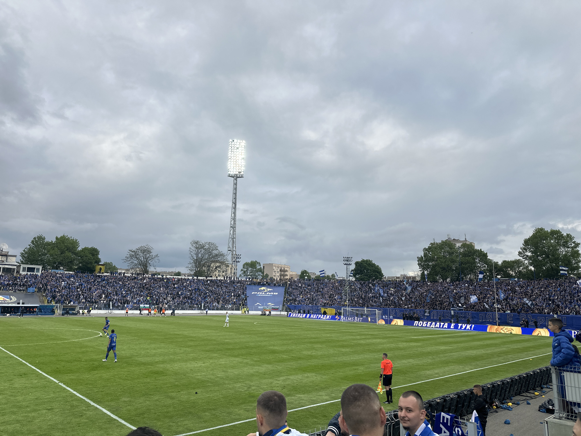 A wide shot captures a soccer match in progress at a stadium in Sofia, Bulgaria. The sky is overcast with a dense layer of gray clouds. In the center of the frame is a tall, white stadium light tower. The stadium is packed with spectators, primarily dressed in blue, filling the stands on both sides. Players in blue uniforms are visible on the green soccer field, while a referee in an orange uniform stands near the sideline. Banners with text in Bulgarian, including phrases like "ХИЛЯДИ В НАГРАДИ! PALMS BET," are displayed along the stadium perimeter. People are visible in the foreground, suggesting the viewpoint is from within the stadium. Trees surround the stadium, adding to the overall environment. The image focuses on the game, the crowd, and the stadium infrastructure.