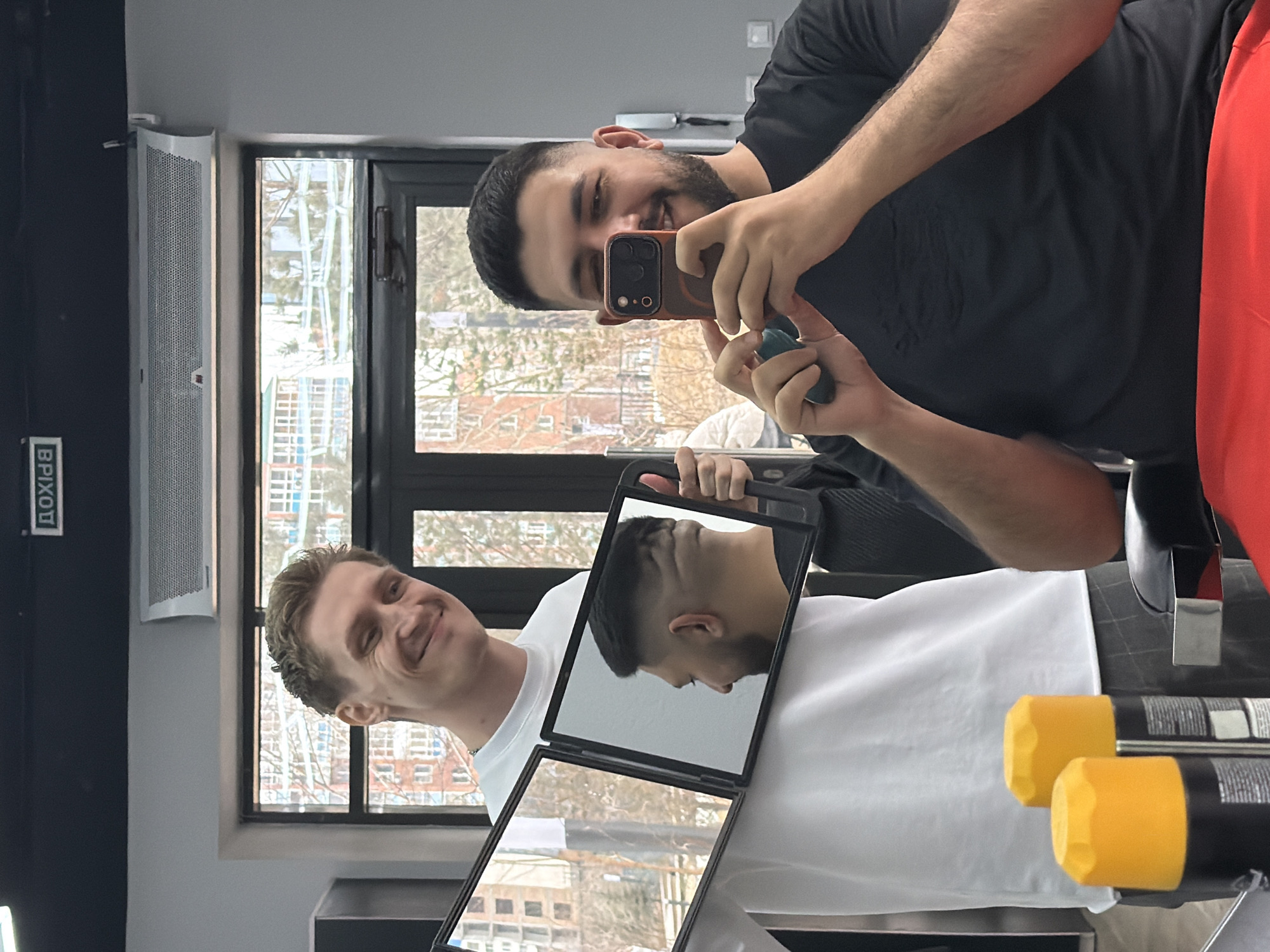 Two men are photographed indoors, one holding a phone and the other a mirror, likely documenting a haircut. The background reveals a cityscape through a window, identifying the location as Krasnoyarsk, Russia, based on the Cyrillic signage visible. One man wears a black shirt and red pants, taking the photo. The other is wearing a white shirt, smiling, and holding a folding mirror to display the haircut. Two hair product bottles with yellow lids are visible on a surface near the mirror.