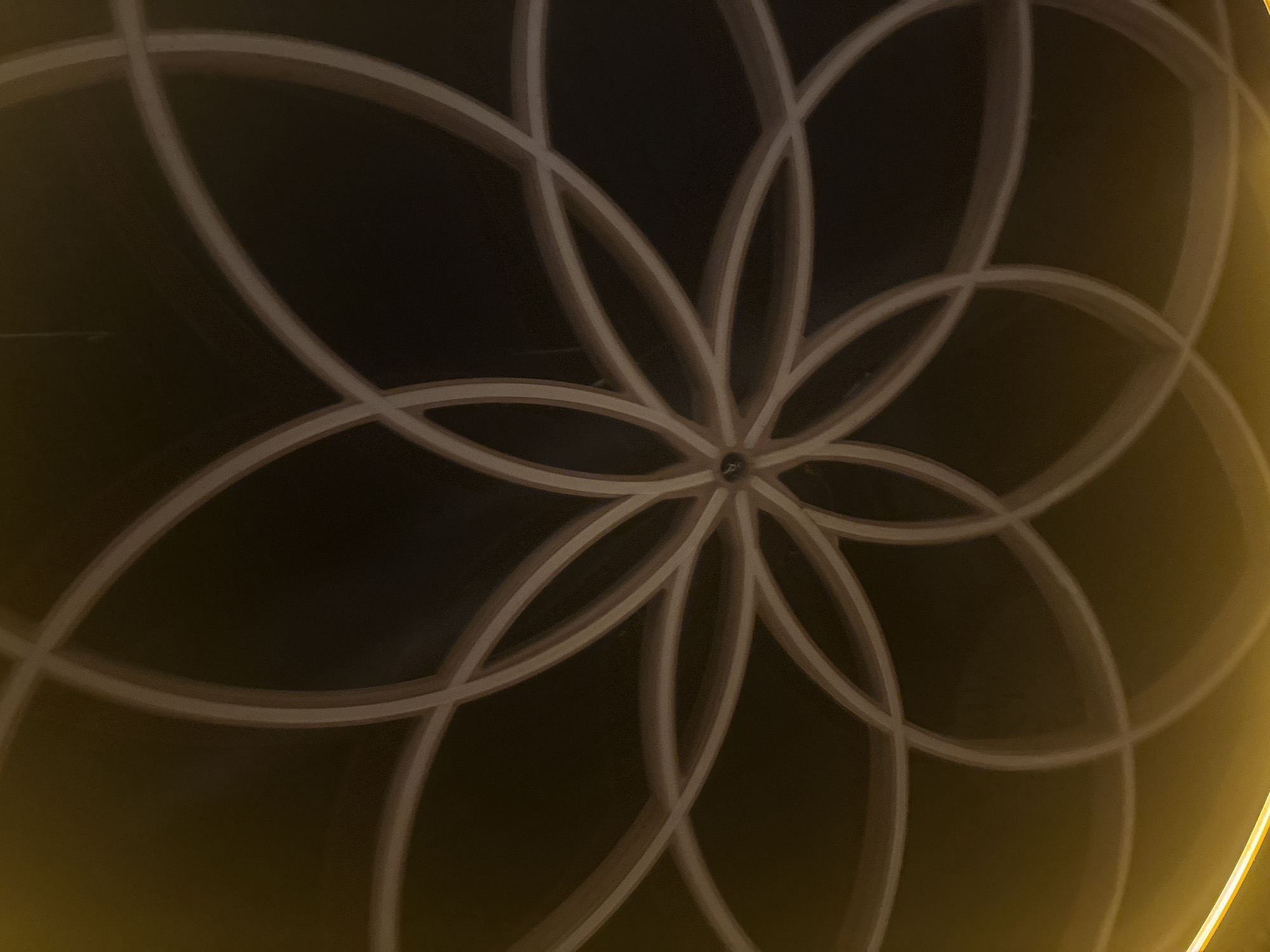 The image shows a close-up of a ceiling with a geometric pattern of overlapping circles. The pattern is illuminated, with a gradient of light from yellow on the left to dark brown in the center and on the right. The circles are light beige and form a symmetrical, flower-like design with eight petals. At the very center of the pattern, there appears to be a small, dark fixture, possibly a light or a decorative element. The overall impression is one of abstract architectural design. There are no people, animals, or specific landmarks visible in the image. Without additional context, it is impossible to determine the exact location within Burgas, Bulgaria.