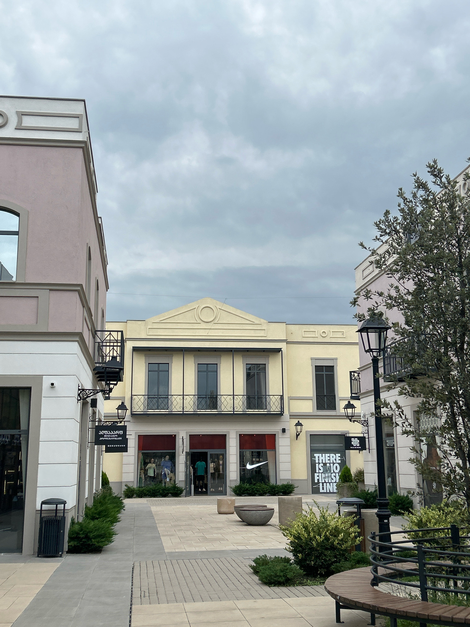 This is an outdoor daytime photograph taken in the city of Gamarjveba, Georgia. The image features a row of commercial buildings with light yellow and pink facades, characteristic of an outdoor shopping complex. In the foreground, a paved walkway with cobblestone accents leads towards the buildings. Several well-maintained green bushes and small trees are planted along the walkway. A dark grey trash receptacle is visible on the left side of the image.
The central building is painted a light yellow and has a neoclassical architectural style with a triangular pediment. It features multiple windows with dark frames and ornate wrought-iron balconies on the upper level. The ground floor of this building houses retail spaces. One storefront prominently displays the Nike logo, and another features a sign with the text "THERE IS NO FINISH LINE" in large white letters.
To the left of the yellow building is a pink structure with white architectural details, also featuring large windows and balconies. To the right, another pink building with white accents is visible, with a tree with dense green foliage partially obscuring it. Black lampposts with a traditional design are positioned along the walkway.
In the middle ground, two men are visible near the entrance of one of the stores, one wearing a green t-shirt and the other a blue t-shirt and shorts. They appear to be employees or shoppers. A black sign with white text, written in Georgian script, is hung on the pink building on the left. Two rounded concrete planters are also present in the shopping area.
The sky is overcast with a grey, cloudy appearance, suggesting a possibly humid or cool day. The overall scene depicts a modern retail environment within a Georgian city.