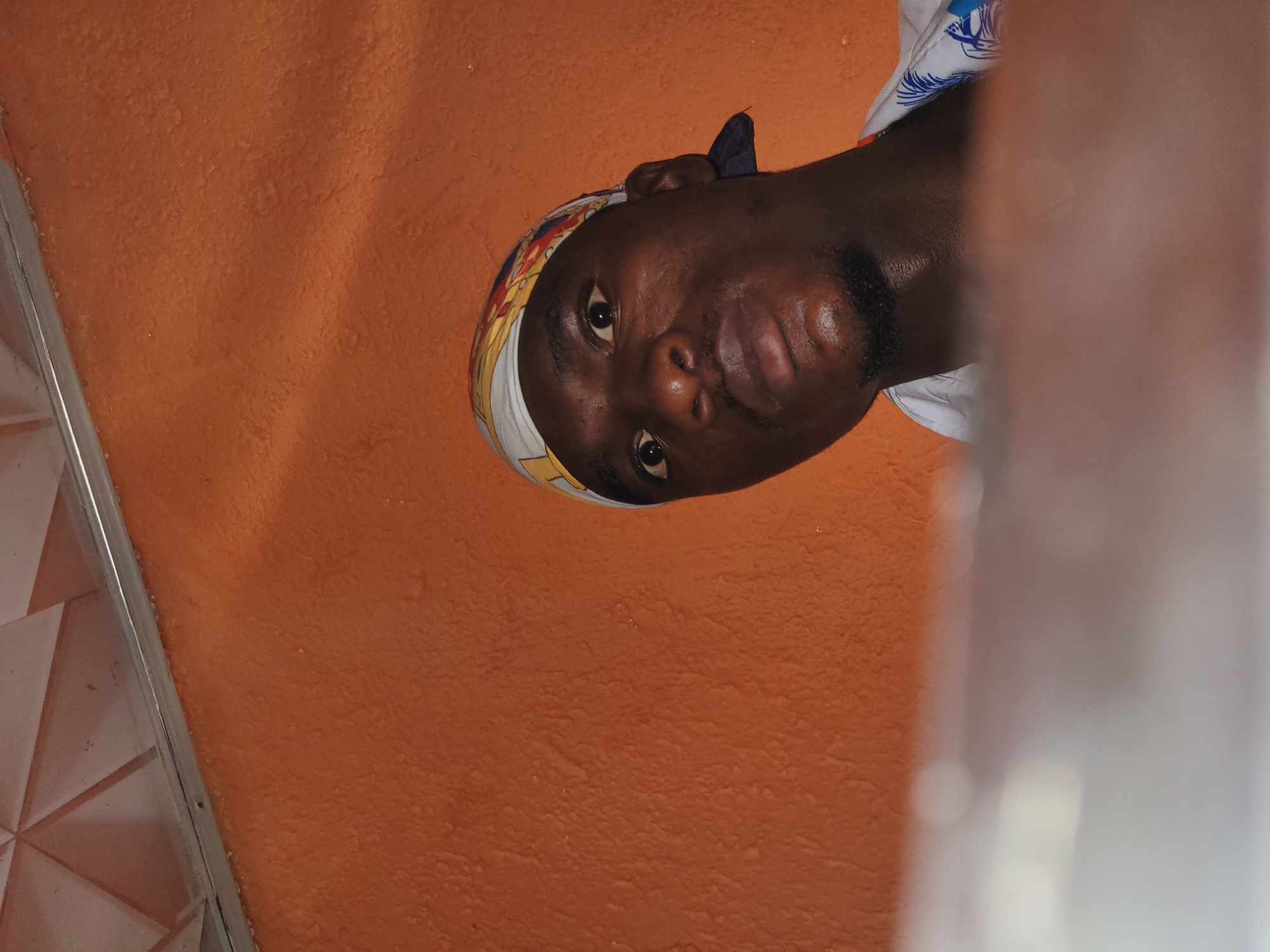 A man wearing a bandana and a white shirt with a blue floral pattern looks directly at the camera from a low angle, his face filling most of the frame. The background is an orange textured wall and a white ceiling with a geometric pattern. A blurry, reflective surface occupies the bottom of the image. The scene appears to be indoors, possibly in a residential area of Bukuru, Nigeria.