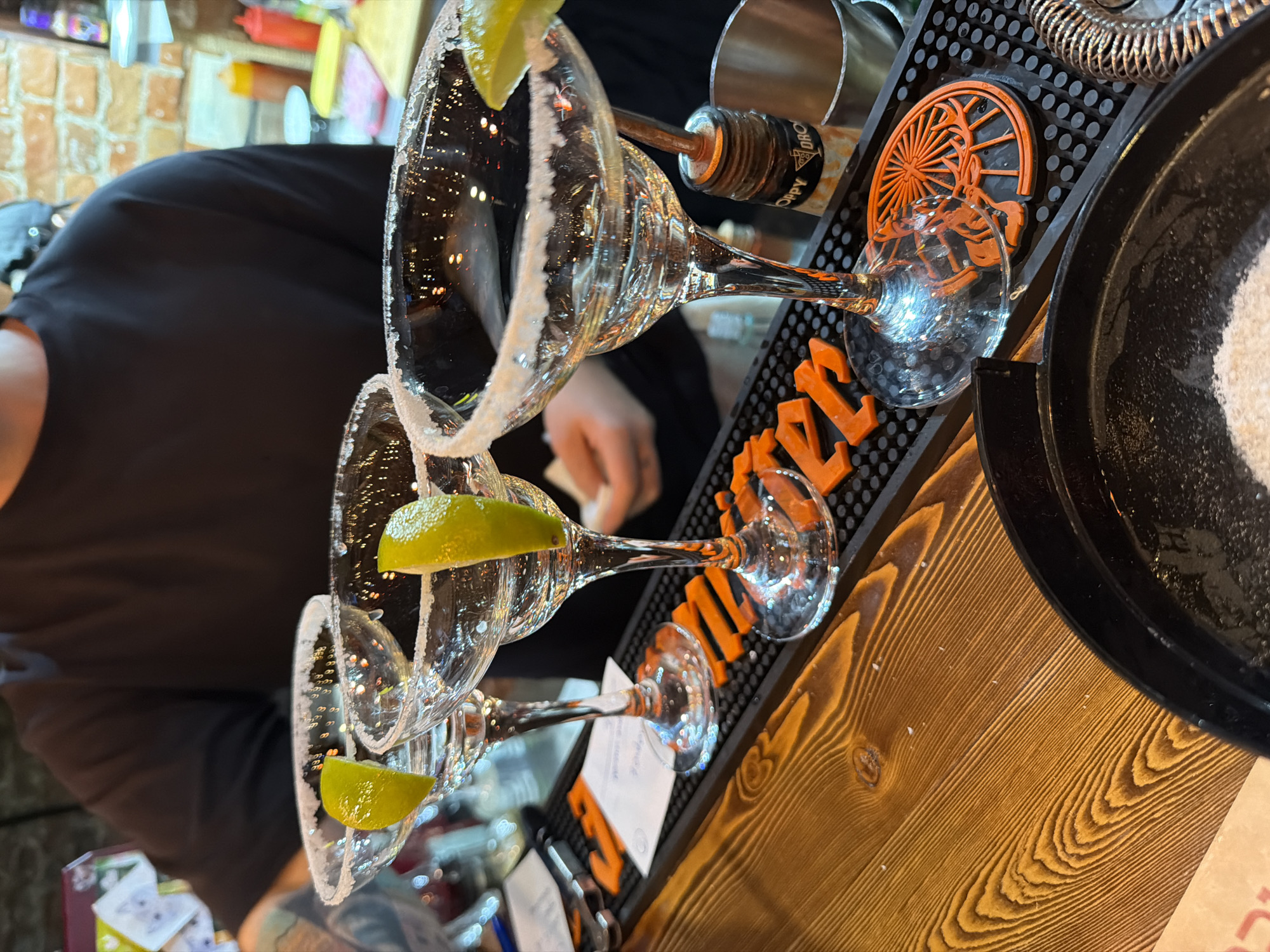 The image shows a bar setup, likely inside a restaurant or bar. Several margarita glasses with salted rims and lime wedges are arranged on a black bar mat with orange accents. The brand name "Jägermeister" is visible on the mat. A speed pourer is attached to a liquor bottle next to the glasses. Part of a person wearing a black shirt is visible in the background, potentially preparing drinks. A salt rimming tray is situated to the right. The bar top is made of light-colored wood. The image might be taken in Krasnoyarsk, Russia, but there is no explicit indication within the image itself.