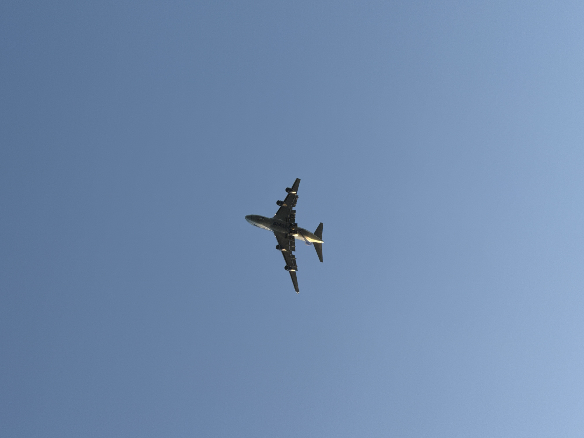 A large, dark aircraft is captured flying across a clear, bright blue sky, viewed from directly below. The plane, likely a wide-body jet such as a Boeing 747, is distinguishable by its four engines mounted under the wings. Its underside is primarily visible, with some reflective highlights on the right wing and tail surfaces, suggesting sunlight from above and to the right. The landing gear is retracted, indicating it is in flight. The sky is uniform and cloudless, signifying clear weather conditions during the daytime. On the underside of the fuselage, partially legible text can be observed, possibly reading "NATIONAL" or a similar airline name. The aircraft appears to be moving away from the viewer, receding into the distance. This scene takes place in the sky above Tel-Aviv, Israel.