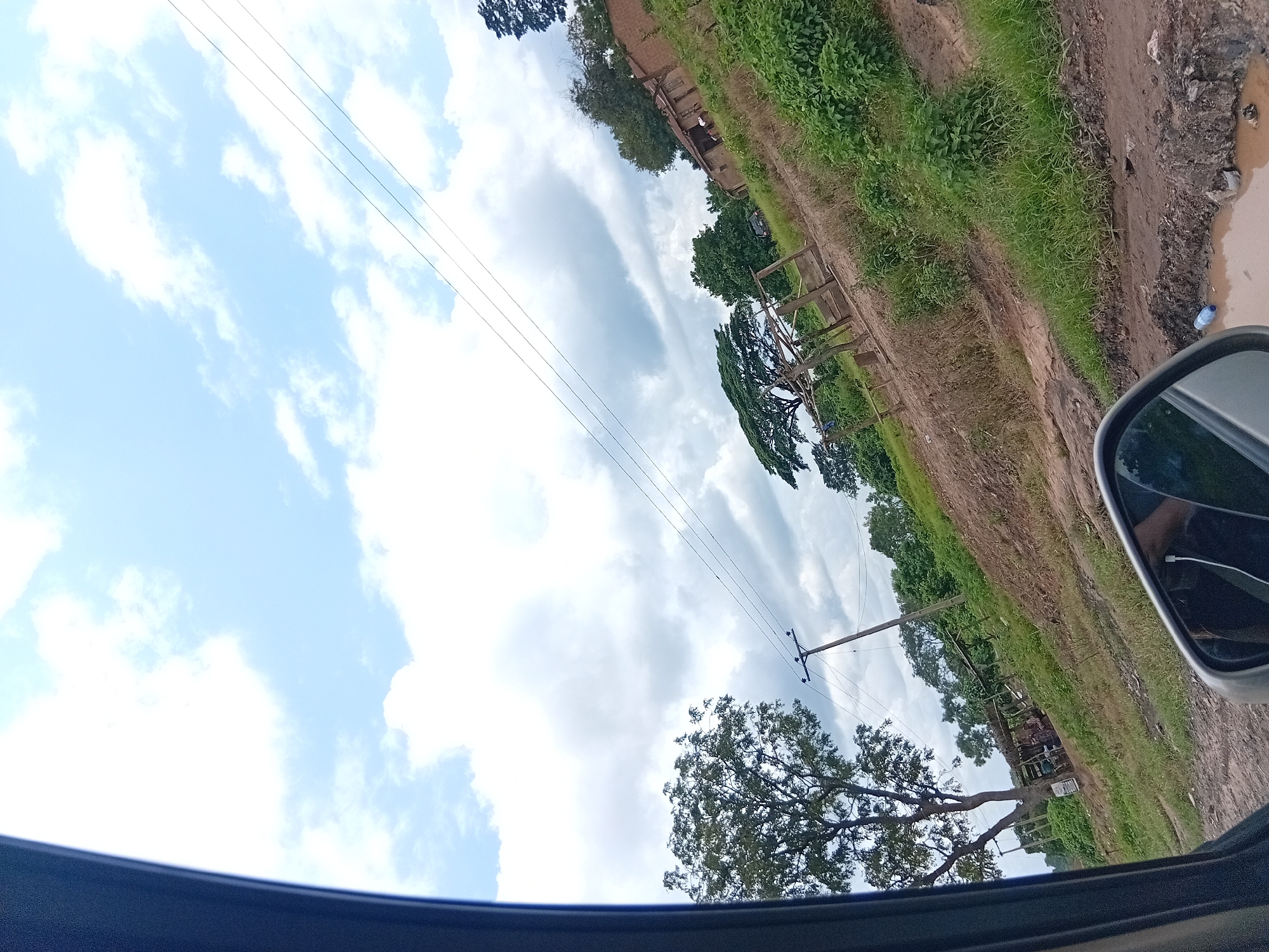 The image depicts a rural roadside scene in Adoma, Nigeria. A dirt road, rutted and muddy in places with a puddle reflecting the sky, runs alongside a grassy embankment dotted with bushes and trees. In the middle ground, there is a structure that appears to be a small dwelling or animal enclosure constructed with mud bricks and a thatched roof, with some people visible underneath. Further back, a larger tree with a wide canopy stands near a collection of similar structures. Power lines stretch across the sky, supported by a wooden utility pole. The sky is partly cloudy, with a mix of white cumulus clouds against a pale blue backdrop. A car's side mirror is visible in the foreground, indicating the perspective is from inside a vehicle.