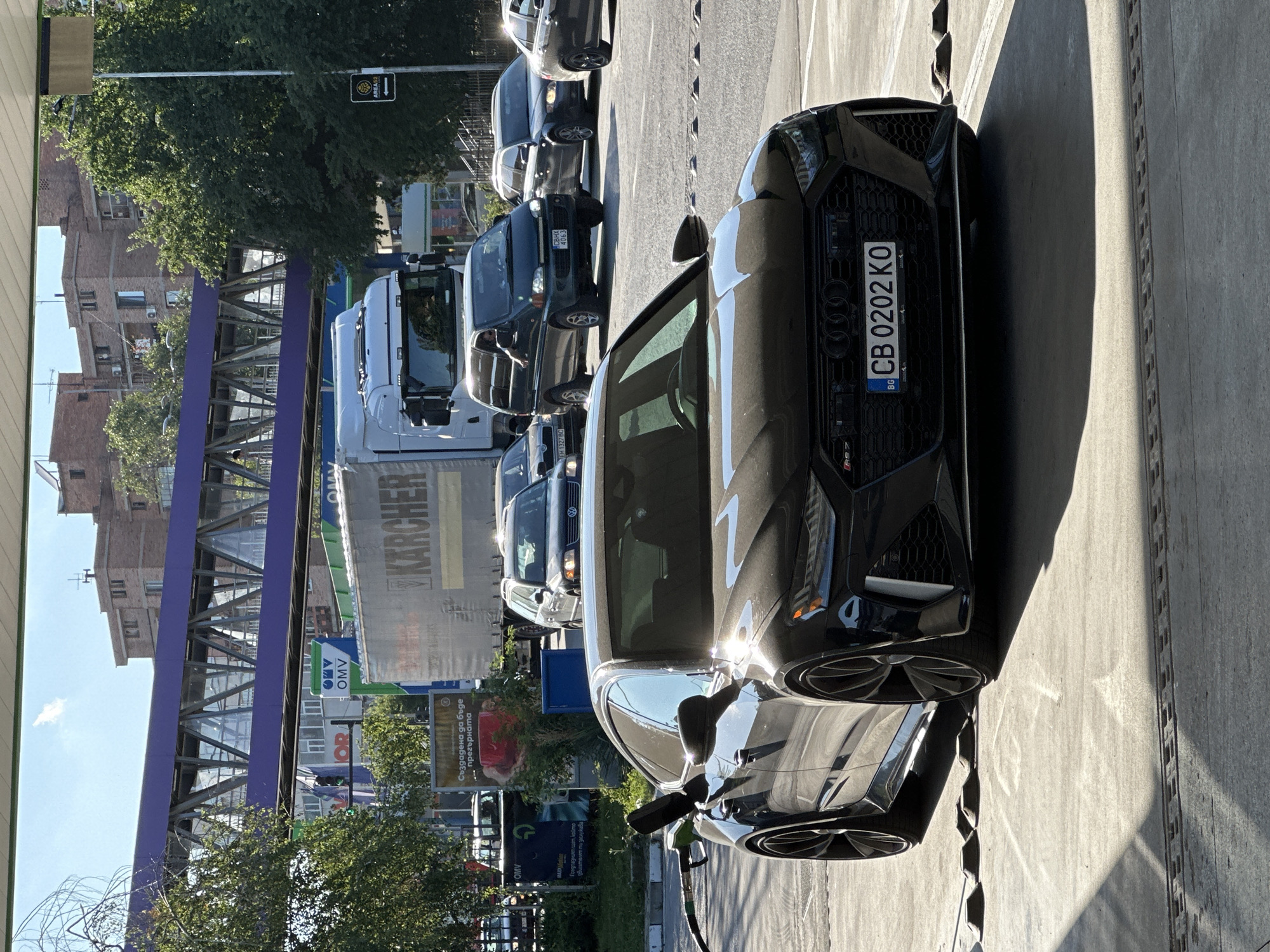 An Audi RS7 is parked at a gas station on a sunny day, likely in Veliko Tarnovo, Bulgaria, as indicated by the license plate format "BG CB 0202 KO". The car is black and positioned at an angle facing the camera, with the driver's side door open and a green fuel nozzle inserted. The background features a pedestrian bridge with purple accents, a white Karcher-branded truck, and several other vehicles on a road. Brick buildings and trees are visible in the distance. The ground is paved concrete with tire stops in front of the car.