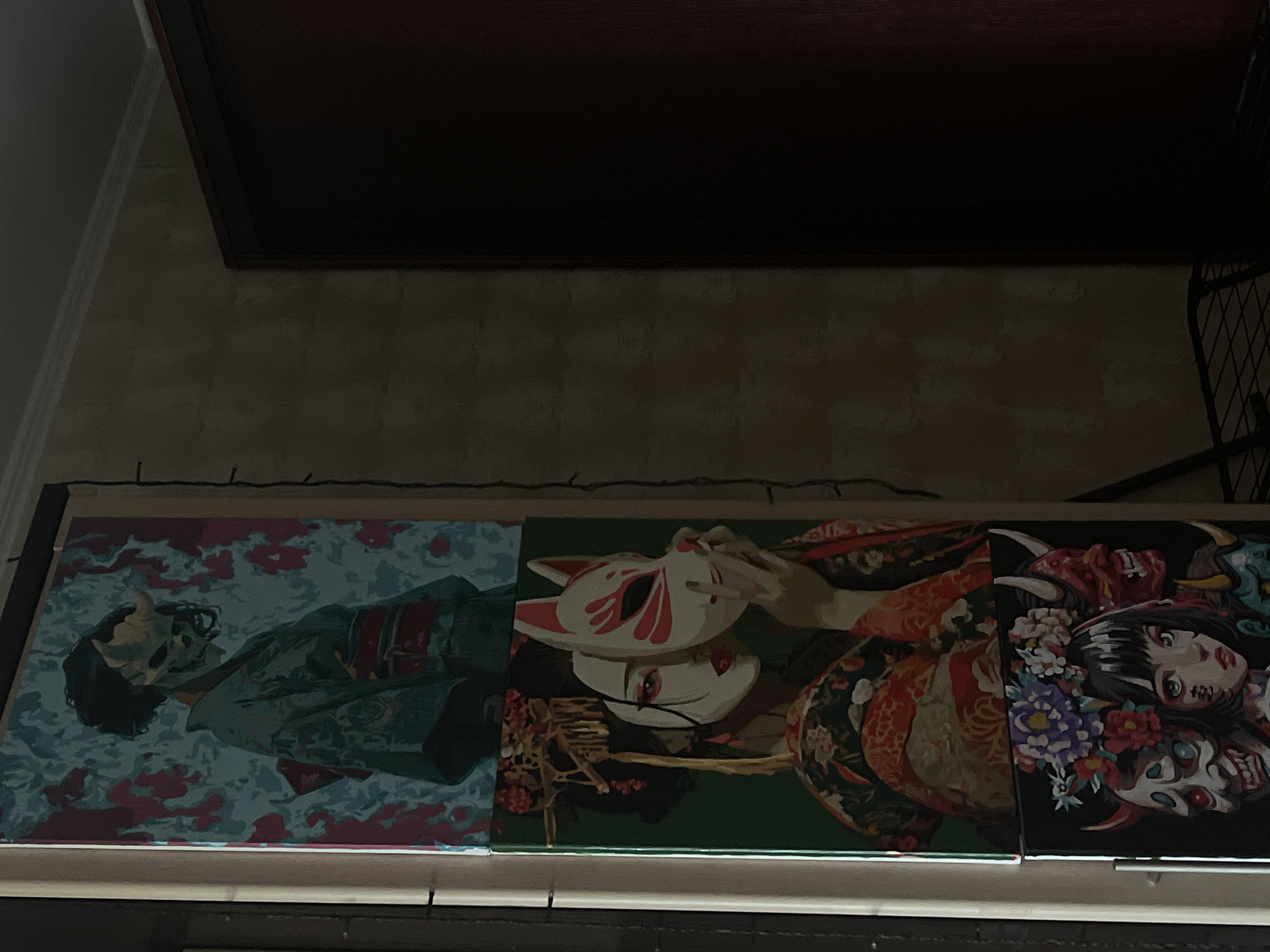 Three vertical canvases are stacked and displayed on a light-colored wall, possibly a door frame. The top canvas depicts a figure in a kimono wearing a horned, skull-like mask against a teal and pink floral background. The middle canvas shows a woman in a traditional kimono holding a white fox mask with red accents to her face. The bottom canvas features a young woman with a flower crown, surrounded by three demonic masks. The canvases are positioned to the left of a dark, vertical surface, possibly a wardrobe or door, and to the right of a wall with a textured pattern. A string of small lights hangs down the left side of the wall. The overall lighting is dim.