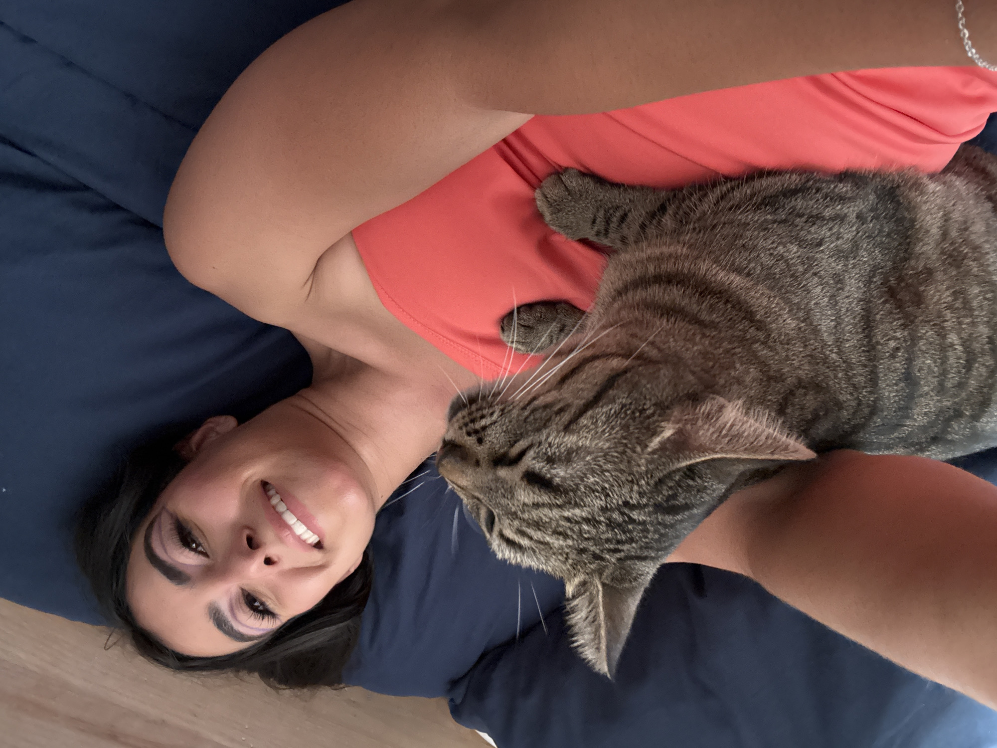 A woman lies on her back on a blue bed, smiling. She wears a coral-colored tank top. A tabby cat rests on her chest and arm. The location is unknown; there is no evidence suggesting Puebla, Mexico.