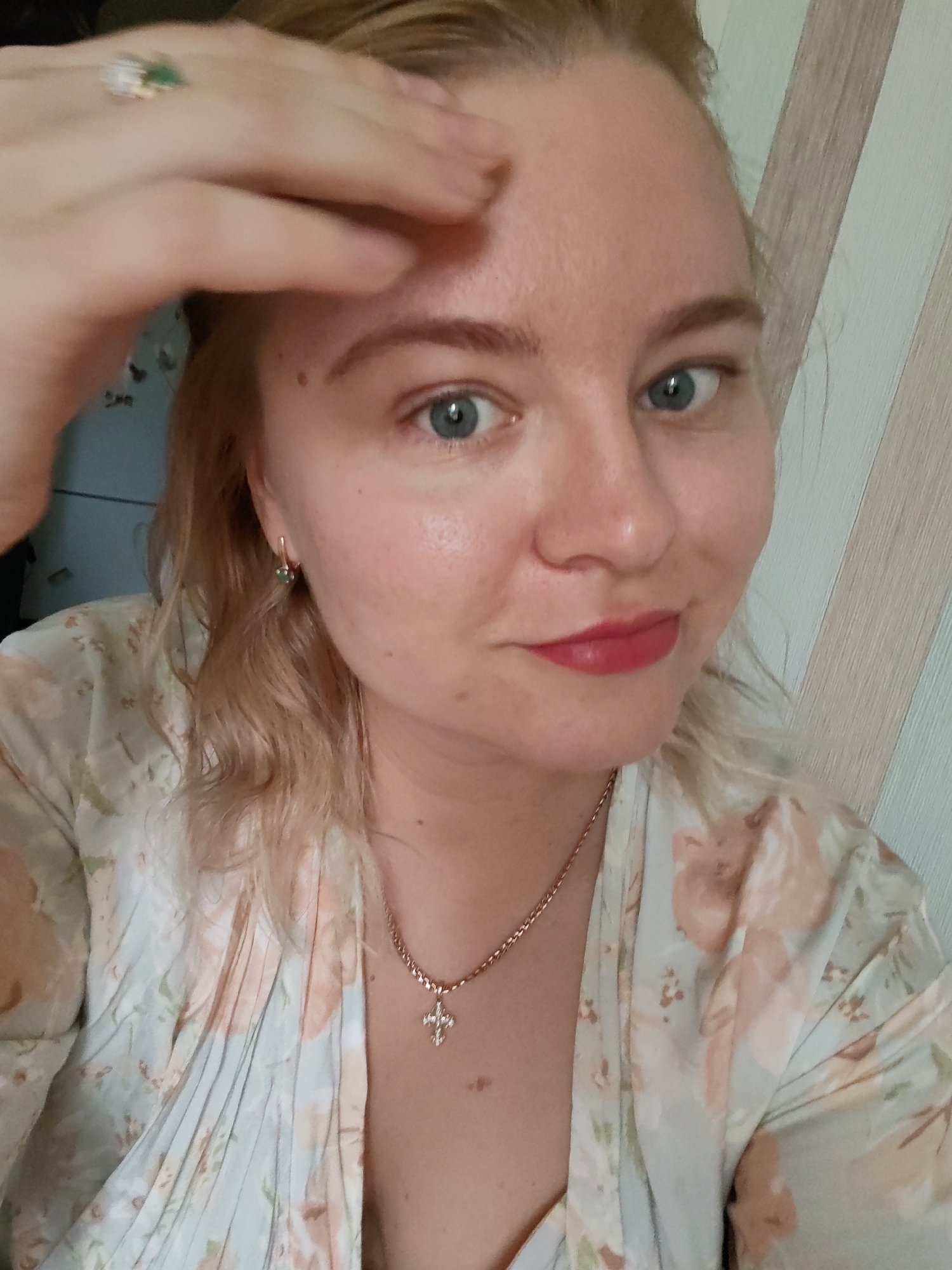 A fair-skinned woman is the focus of a close-up, selfie-style photograph. She has light blond hair, visible blue eyes, and is wearing red lipstick. Her left hand is raised to her forehead. She's wearing a ring on her left hand and earrings. A thin gold chain necklace with a cross pendant is visible around her neck. She's wearing a floral patterned garment. In the background, there is a light blue wall on the left and a beige and white striped wall on the right. The photograph does not contain specific details indicating the location is Izhevsk, Russia.
