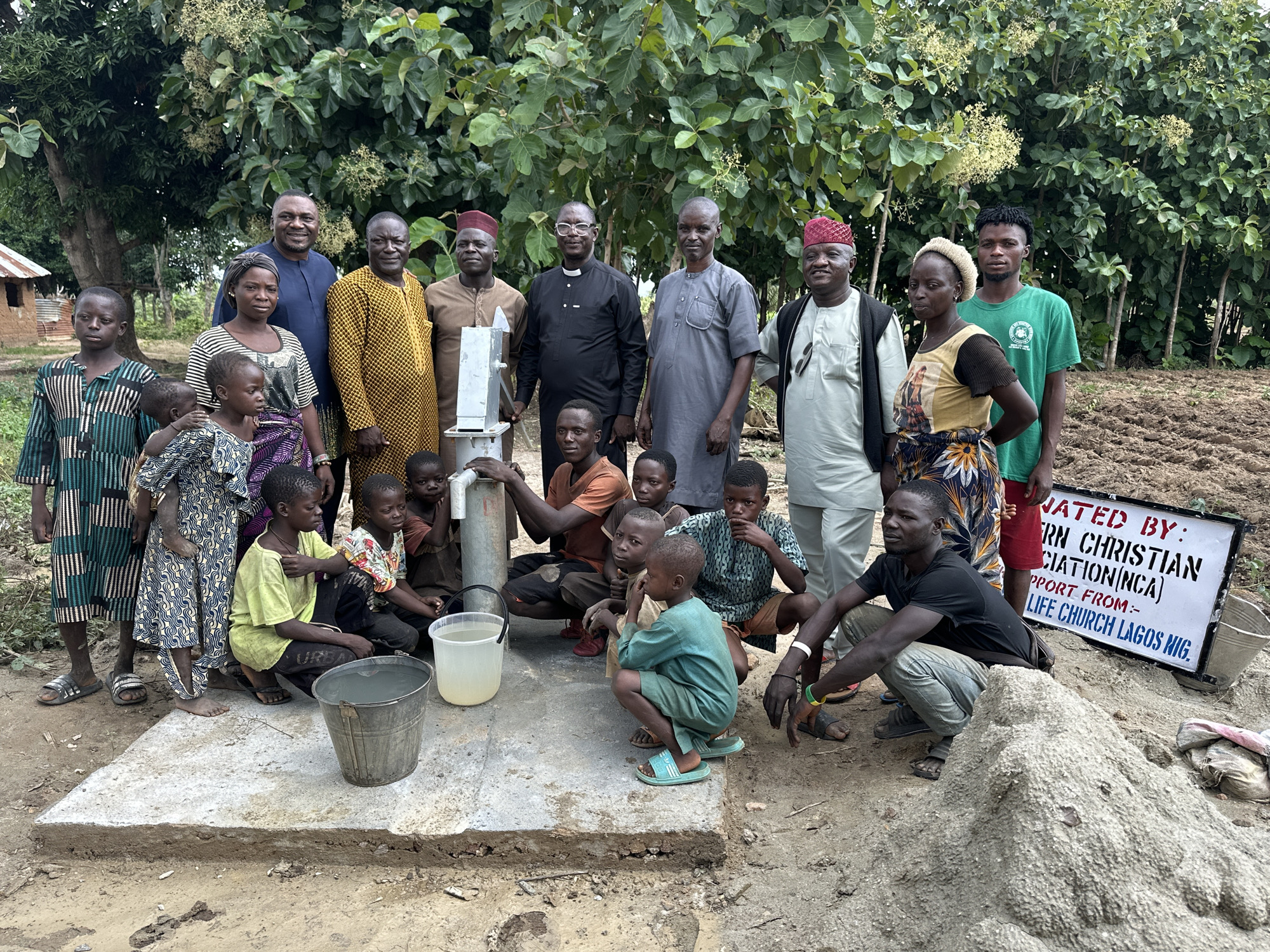 In Afogo, Nigeria, a group of approximately twenty people, comprising both adults and children, are gathered outdoors around a newly constructed water well. The individuals are of varying ages and genders, and many of them are dressed in traditional African attire. The scene appears to be a community event, possibly a dedication or unveiling of the well. The environment is rural, with a backdrop of trees and vegetation, and the ground is composed of dirt and some loose soil. A sign is visible in the background, which reads "RN CHRISTIAN ASSOCIATION (NCA) SUPPORT FROM:- LIFE CHURCH LAGOS NIG.". The people are interacting with each other in a relaxed and friendly manner, with some of the children playing near the well. The overall atmosphere is one of communal activity and shared purpose.