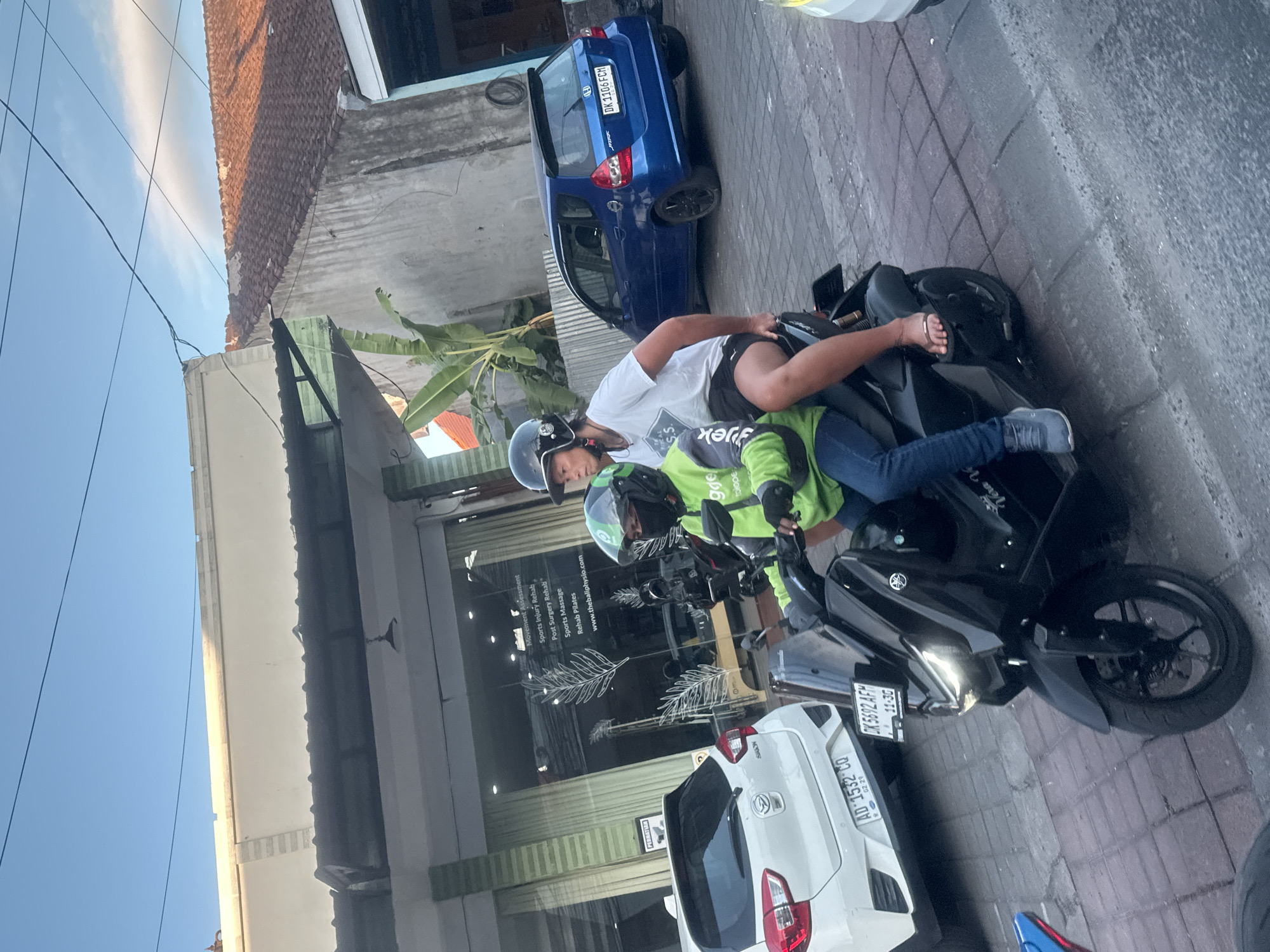 In North Kuta, Indonesia, two individuals are seen riding a black motorcycle. The person in the front, wearing a green jacket and a black helmet with a face mask, is operating the vehicle. The passenger behind, wearing a white t-shirt and black shorts, has their legs exposed. The scene appears to be on a street with buildings and vehicles present. A white car is parked to the left, and a blue car is visible further down the road. The sky is blue with some clouds, and the lighting suggests it is daytime. The road is paved with bricks. The motorcycle has a visible license plate.