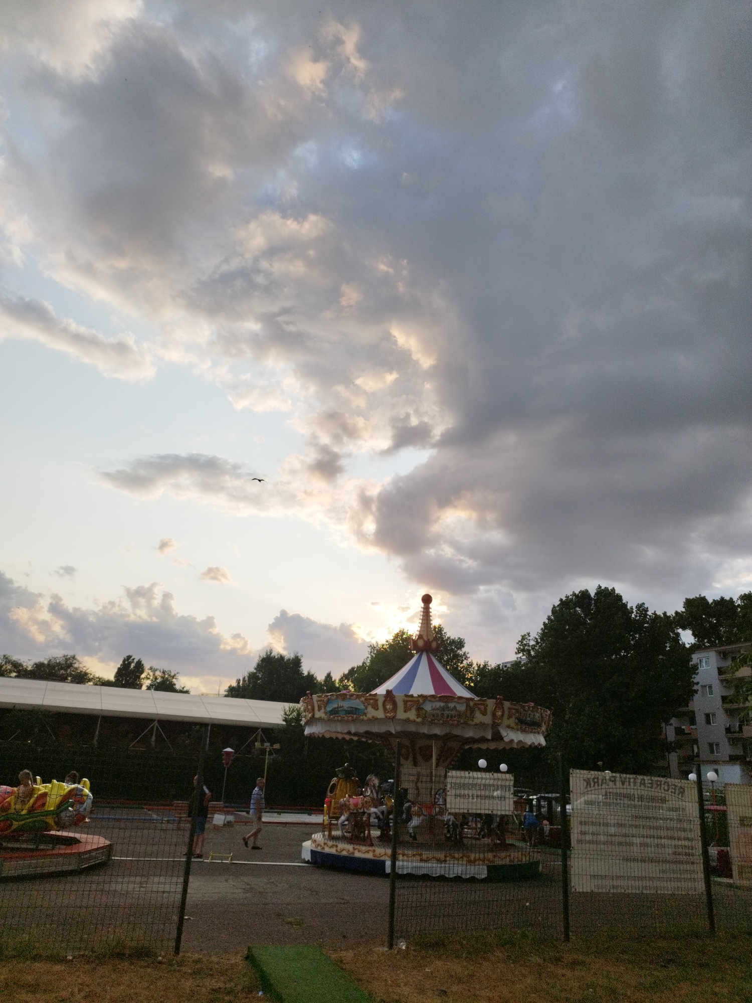 The image captures a scene at "Recreativ Park" in Constanța, Romania, during the late afternoon or early evening. The sky is filled with dramatic, dark clouds, with patches of lighter sky and sunlight breaking through, suggesting a transition in weather. In the foreground, a black chain-link fence encloses a park area. Beyond the fence, a carousel with a red, white, and blue striped canopy is visible, featuring horses and other figures. To the left of the carousel, a smaller, brightly colored amusement ride with yellow and red cars is also present, with a child seated in one of the cars. Several adults are scattered throughout the park, some walking and others standing near the rides. In the background, a long, white tent-like structure is set up, and behind it, dense green trees frame the scene. A multi-story building with balconies is visible on the far right, partially obscured by trees. Two informational signs, one with text that reads "RECREATIV PARK" in reverse, are positioned behind the fence. The ground within the park is paved, with areas of dry grass and some green turf visible along the fence line. A lone bird is silhouetted against the sky in the upper left quadrant of the image.