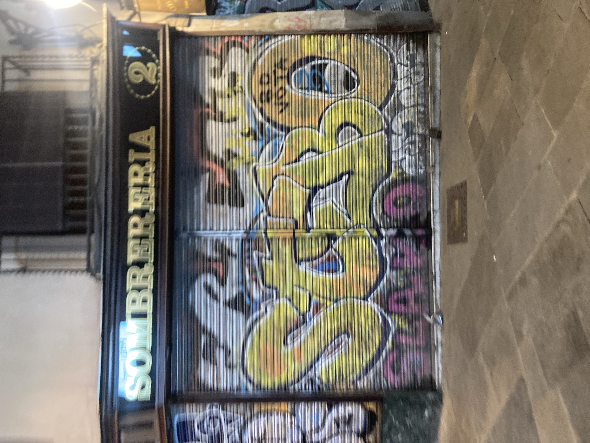 A storefront in Barcelona, Spain, is depicted with a neon sign reading "SOMBRERERIA" in gold lettering and a circular emblem containing the number "2". The storefront's metal rolling shutters are covered in graffiti. Large, stylized letters in yellow and orange dominate the shutters, with visible outlines and shading. Some of the graffiti includes what appears to be the words "STAR" and "VOTA" in smaller text within the larger designs. Below the main graffiti, a line of pink and purple text is visible, though illegible. The street in front of the storefront is paved with stone tiles, and a doormat is placed near the entrance. The scene appears to be at night or in low light conditions, with artificial lighting illuminating the signage and graffiti.