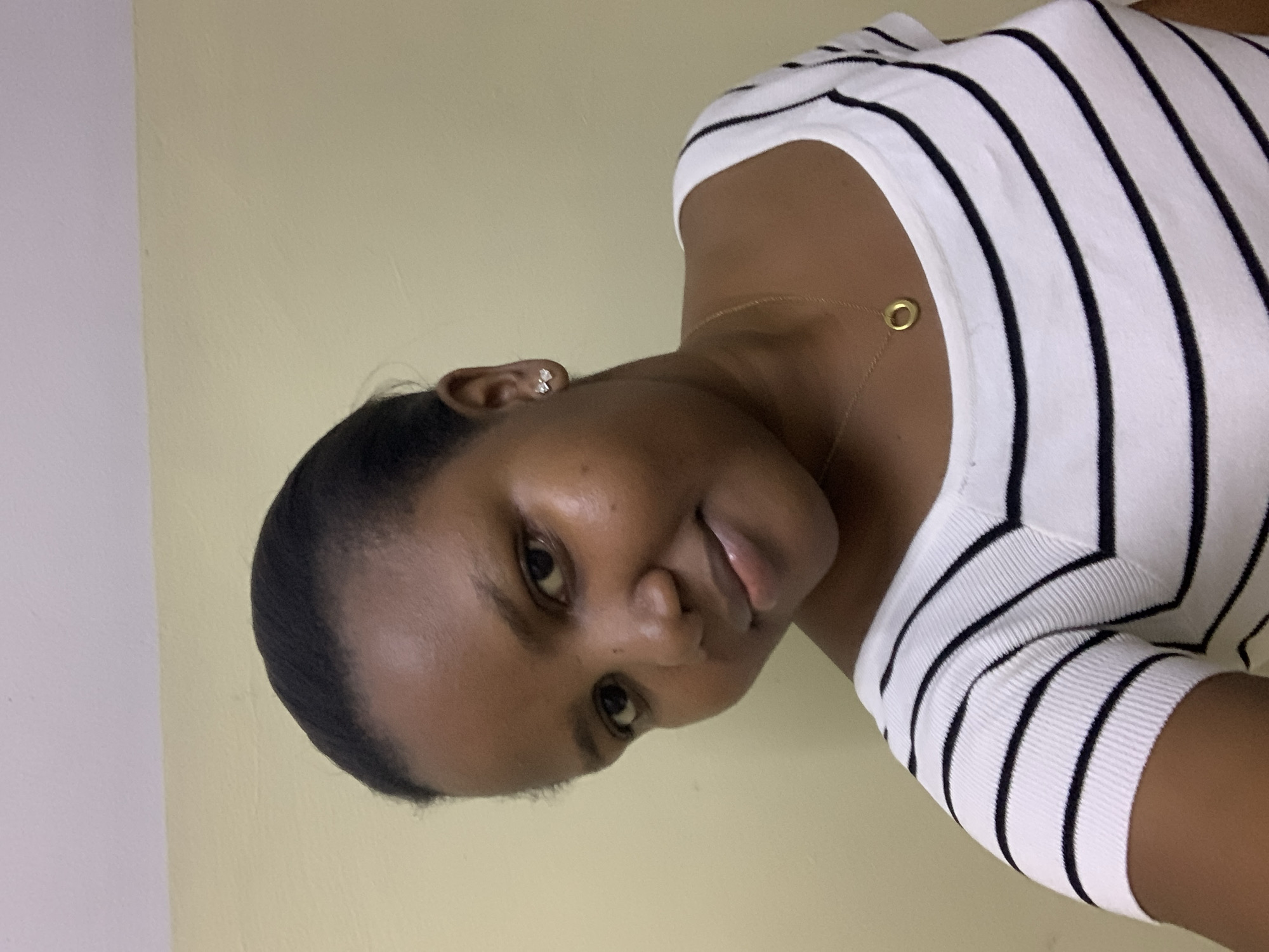 A medium shot of a woman in front of a pale yellow wall. She is wearing a white and black striped shirt and a gold necklace. Her hair is black and pulled back. She has an earring in her left ear. The location is not discernible from the image and no information suggests the city of Dar es-Salaam, Tanzania.