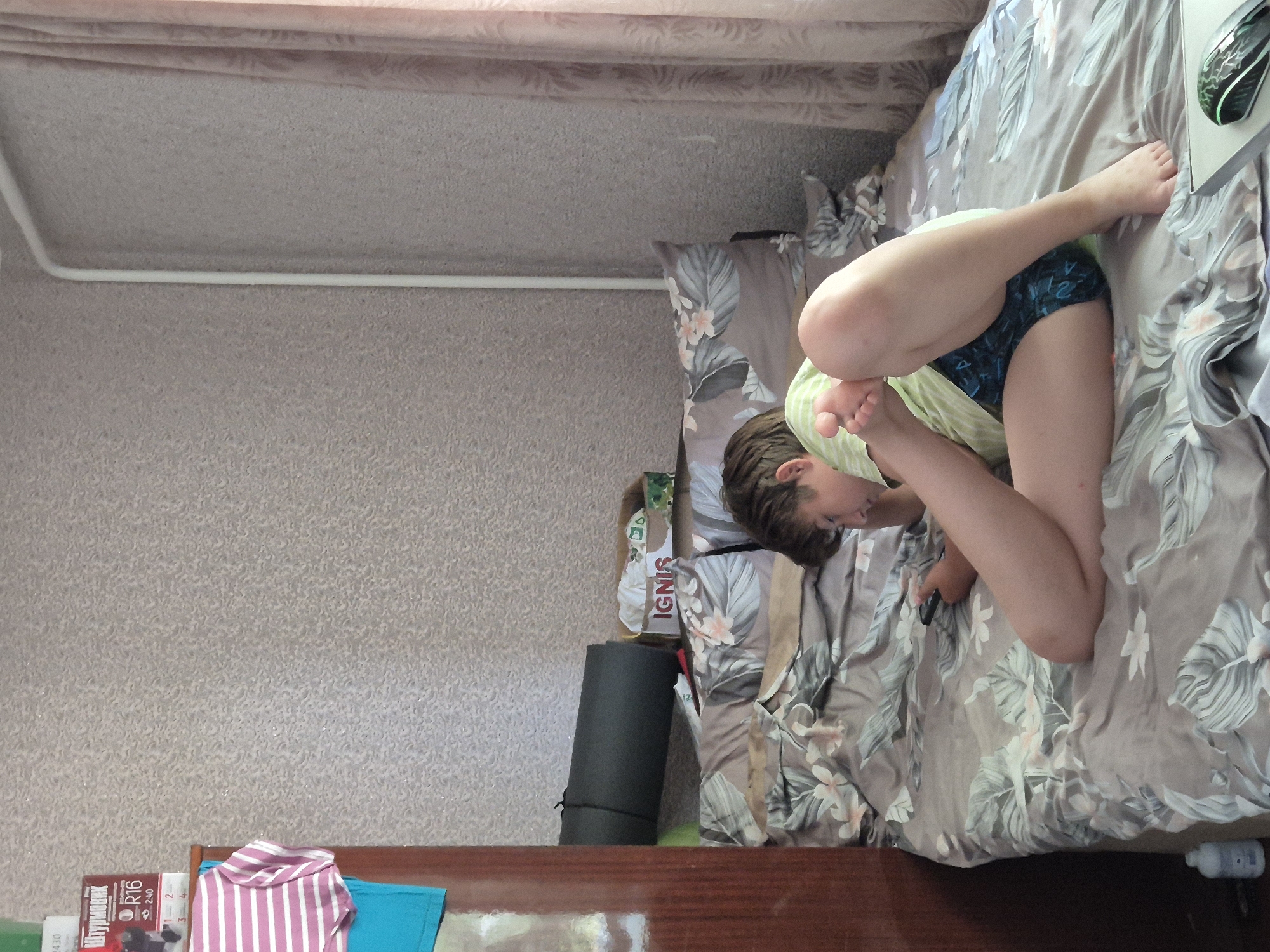 A boy is lying on a bed in a room, playing a video game. The bed has a floral pattern comforter and pillows. The boy is wearing a green t-shirt and blue patterned shorts. He is propped up on his arm, with his legs bent. In the background, there is a dresser with clothes hanging on it and a rolled-up sleeping mat. There is also a box with "IGNIS" written on it. A computer mouse is visible on the right side of the bed. The room has patterned wallpaper and a light-colored curtain. This scene appears to be in a residential setting in Sozonivka, Ukraine.