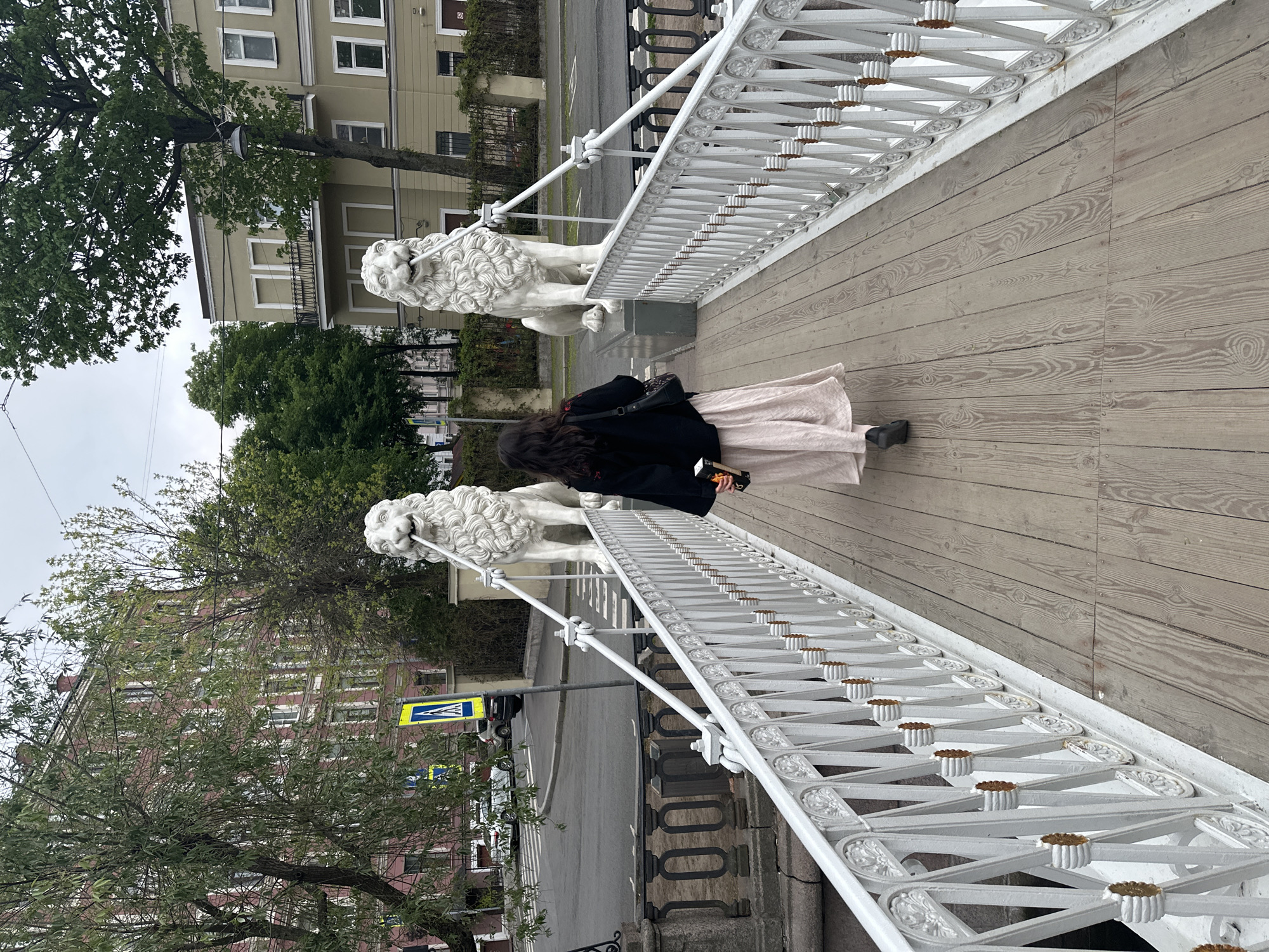 A woman with dark hair, wearing a black jacket and a light pink, flowing skirt, is walking on a wooden bridge with white ornate railings adorned with lion statues. The location is in Saint Petersburg, Russia. Buildings and trees are visible in the background. A street with a pedestrian crossing sign is seen on the left side of the frame.