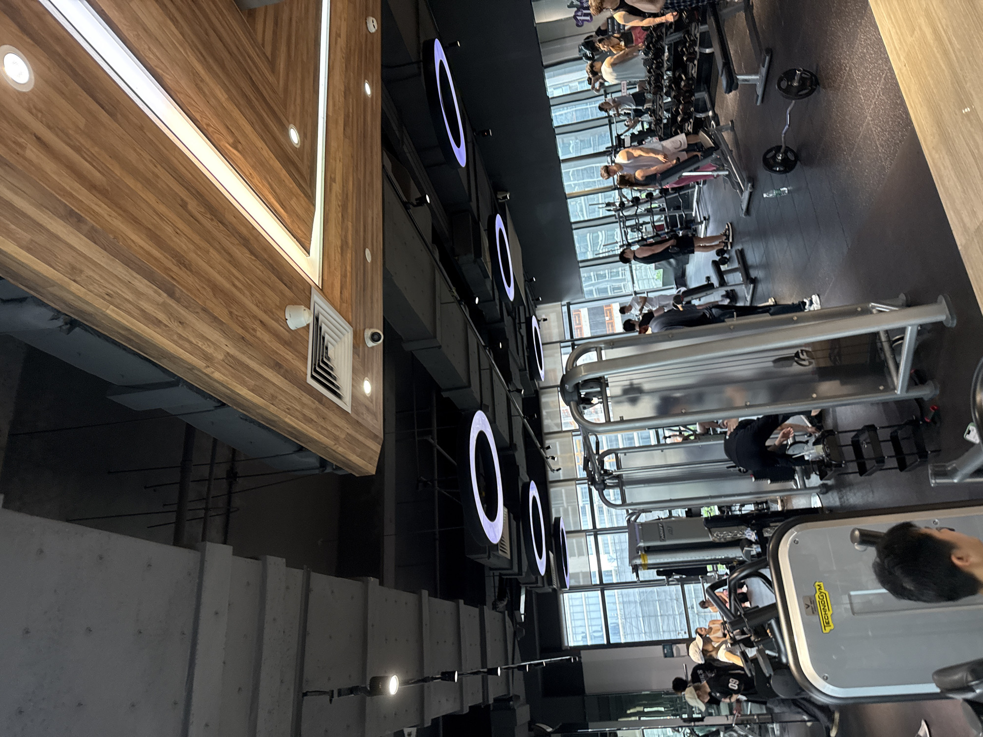 The image depicts the interior of a modern gym in Haizhu District, China. The space features high ceilings with exposed ductwork and industrial lighting fixtures, complemented by wooden accents on the ceiling and walls. Large windows offer a view of urban cityscapes.
In the foreground, a person with short black hair is partially visible, looking away from the camera. To the left, a person wearing a white cap and a black t-shirt with "09" is engaged with a gym machine. A woman in a white top and cap stands nearby.
The midground reveals various gym equipment, including weight machines and benches. Several people are exercising or resting. A man in a black t-shirt and shorts stands near a weight rack. A couple is interacting near a weight bench, with the man wearing a white t-shirt and the woman in a pink outfit. Another person in dark clothing is seen bending over near a weight rack.
The background shows more gym equipment, including a row of weight plates. A bottle of water is on the floor near a barbell. People are scattered throughout the gym, engaged in individual workouts. The overall atmosphere is one of active fitness.