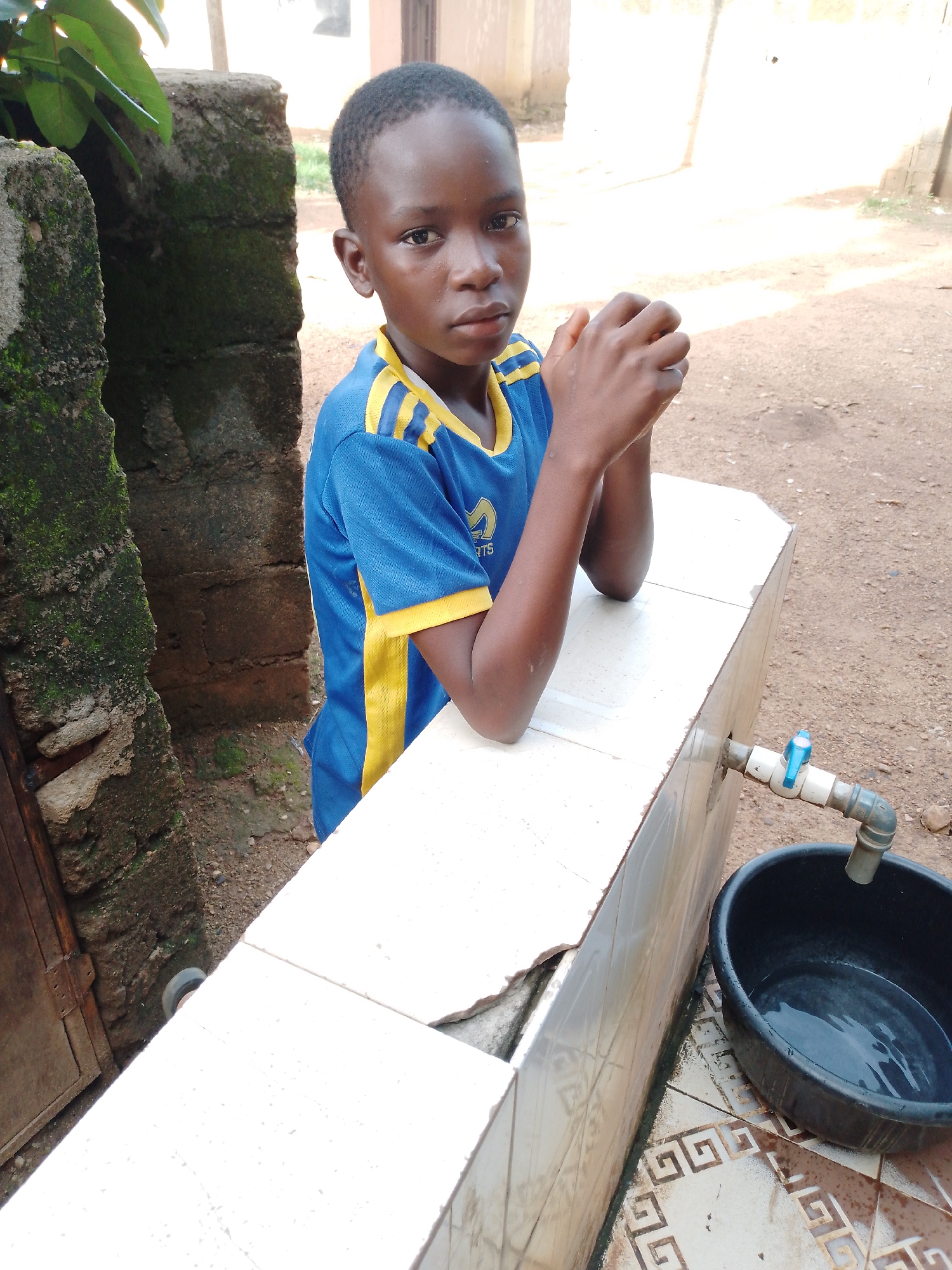 A young boy, likely of African descent, stands leaning on a tiled ledge. He is wearing a blue and yellow t-shirt with a logo that appears to read "RTS". His hands are clasped together in front of him, and he gazes directly at the camera with a neutral expression.
Behind the boy, a rough, moss-covered concrete wall is visible, with some green foliage growing on it. To his right, a black plastic basin filled with water sits on a tiled floor, beneath a pipe with a blue faucet. The scene appears to be outdoors, possibly in a yard or an open-air wash area, given the presence of the faucet and basin. The ground is a mixture of dirt and some patterned tiles. The lighting suggests it is daytime, with bright sunlight casting shadows. There are no other people visible in the frame. The overall impression is one of a simple, everyday moment, possibly related to hygiene or washing.