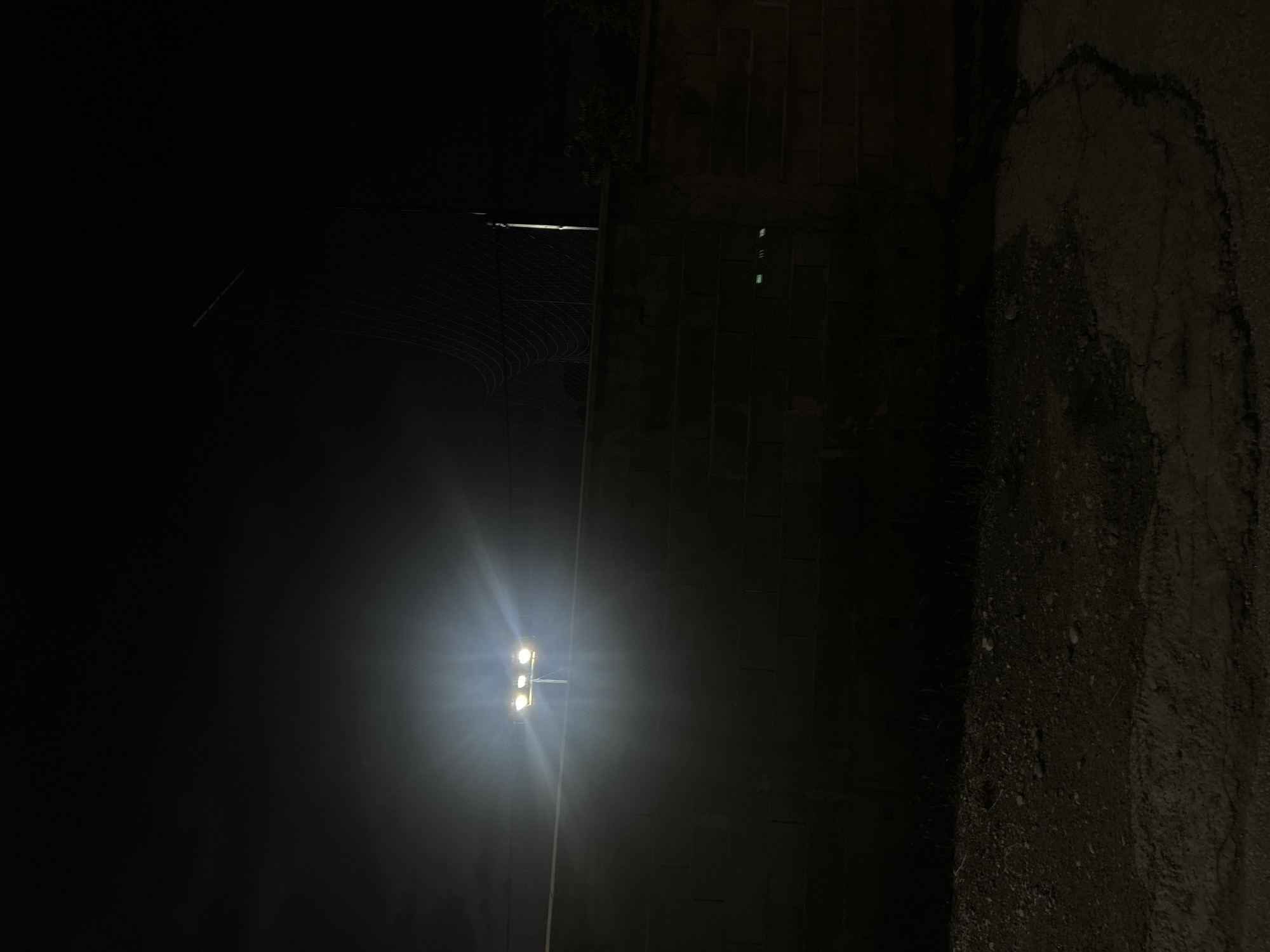 Captured at night, the image presents an outdoor scene, the specific location in the city of Unknown, Unknown, is not discernible. A street or roadway runs vertically along the right side, displaying cracked and weathered asphalt. Along the left is a tall street lamp with three illuminated bulbs. The rest of the image is largely shrouded in darkness, save for the illumination from the street lamp. Overhead electrical wires can be seen in the upper left. The lack of further details prevents a more precise determination of the location or scene's specific characteristics.