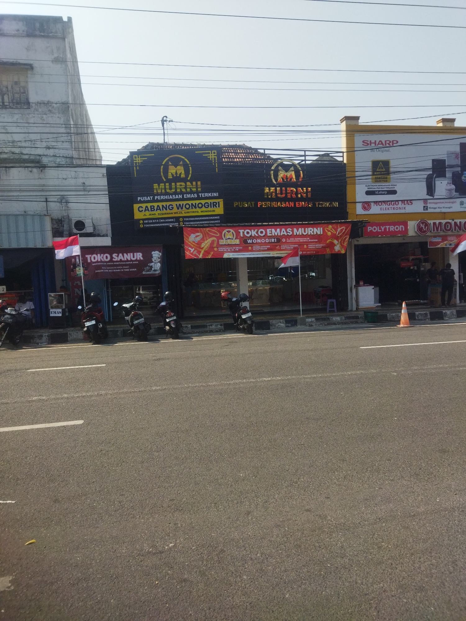The image shows a street scene in front of several storefronts in Wonogiri, Indonesia. The storefronts appear to be a mix of businesses, including a jewelry store with a prominent "MURNI" logo and signage indicating "PUSAT PERHIASAN EMAS TERKINI" (Center for the Latest Gold Jewelry). Another storefront displays "TOKO SANUR" with an image of a chef and text about kitchenware. Further down the street, there's a store with "SHARP" branding and a display of electronic appliances.
Several motorcycles are parked on the sidewalk in front of the businesses, suggesting a mode of transportation common in the area. Indonesian flags are visible, with one prominently displayed in front of the "TOKO SANUR" store. People are seen inside the stores, some appearing to be staff members or customers. The environment is urban, with a paved road and sidewalk, and buildings lining the street. The sky is clear and bright, indicating daylight. The overall impression is of a commercial street in a bustling Indonesian town.