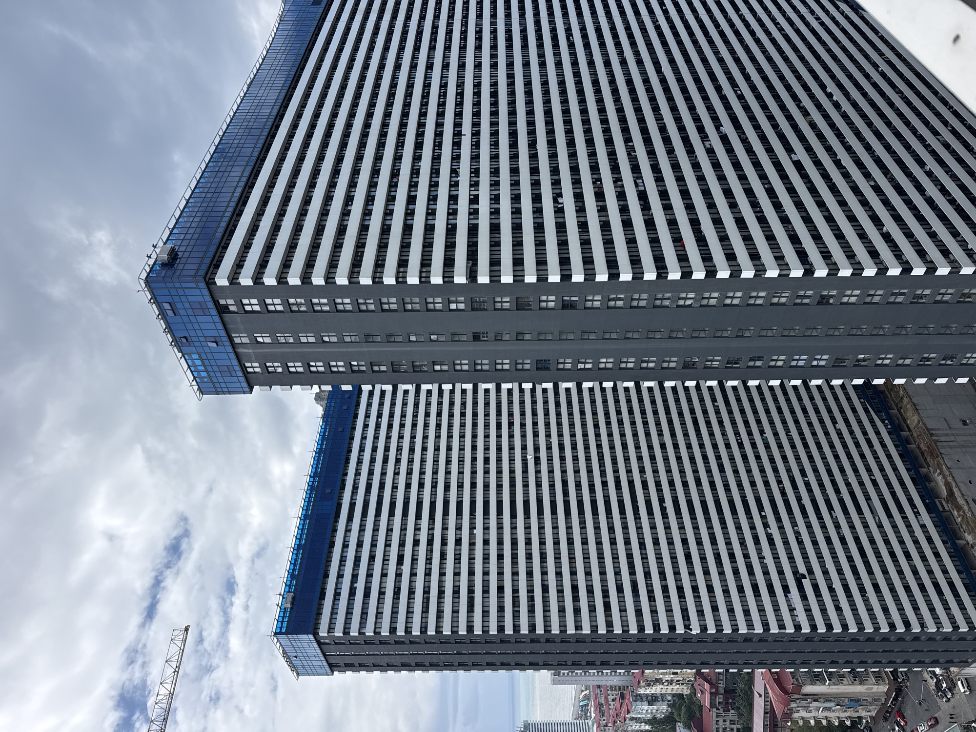 This image, taken in Batumi, Georgia, captures a view of two tall, modern apartment buildings under a cloudy sky. The buildings are characterized by their repeating horizontal white bands and darker grey vertical sections. The tops of both structures are capped with blue glass enclosures, suggesting they may be penthouses or amenities. A construction crane is visible in the upper left corner, indicating ongoing development in the area. Below the main buildings, older, lower-rise structures with red roofs are visible, along with a street with parked cars and a few people. The scene suggests an urban environment that is a mix of new construction and established neighborhoods, with the sea likely nearby given the coastal location of Batumi.