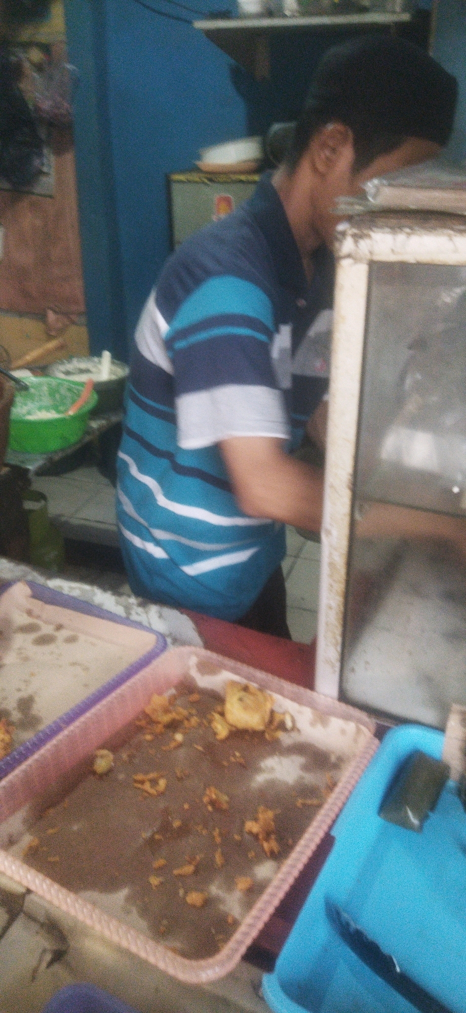 The image captures a close-up, low-angle view of a person wearing a blue and white striped shirt. The person is leaning over a counter with a glass display case in front of them. The person's face is blurred, but they appear to be male. In the foreground, there are two pink plastic trays containing remnants of food. To the right, a blue plastic container holds a dark green item and a pair of black tongs. In the background, a green bowl filled with white powder is visible, along with other kitchen items and a blue wall. The scene suggests a food stall or eatery, possibly located in Depok, Indonesia, given the provided location context.