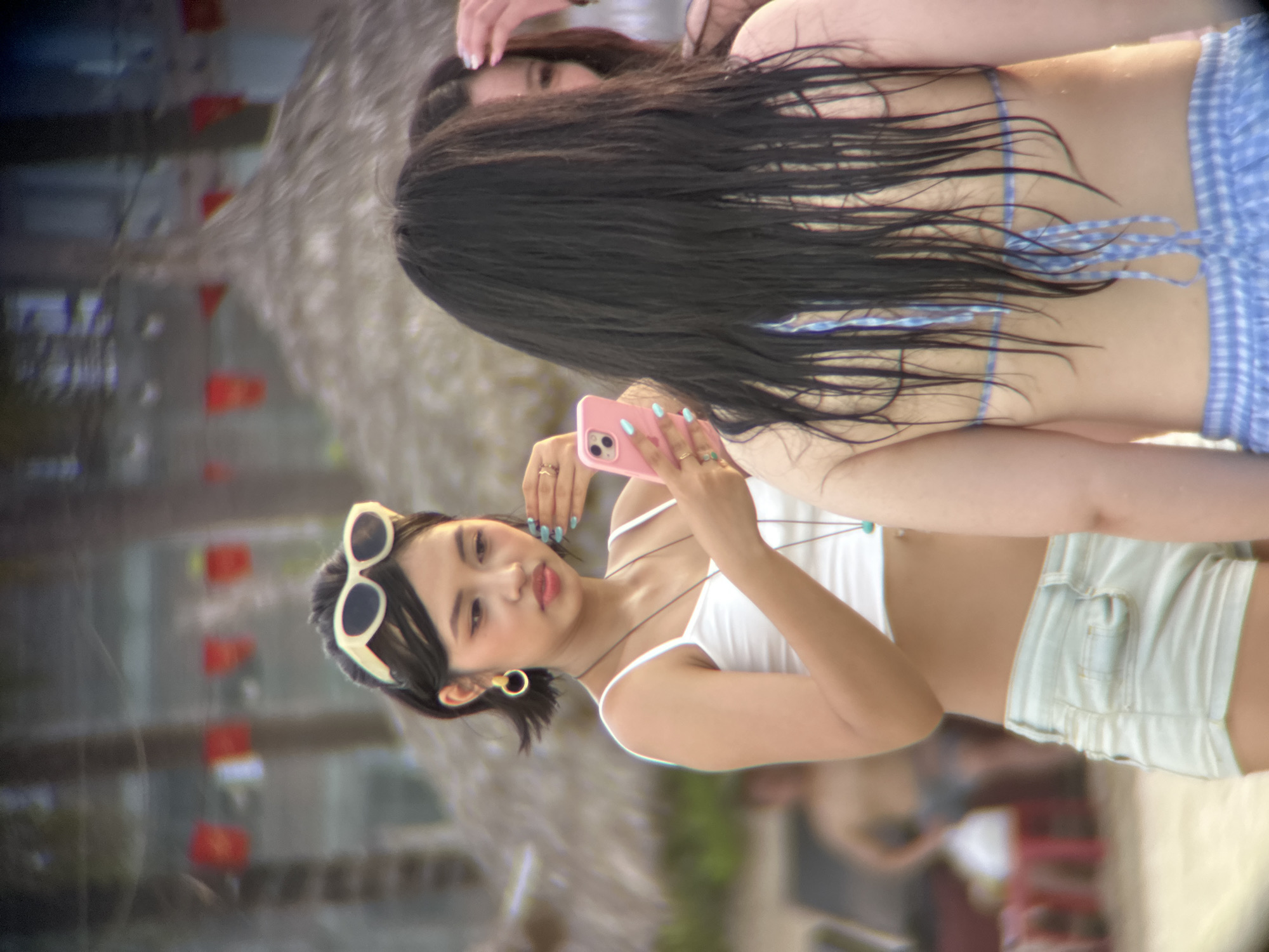 In Đà Nẵng, Vietnam, on a sunny beach, three young women are gathered. The woman in the foreground, on the left, is facing the camera, holding a pink smartphone and taking a picture. She has short, dark hair, is wearing sunglasses, a white crop top, and light blue denim shorts. She is also wearing large hoop earrings and a necklace with a blue pendant. Another woman, whose back is to the camera, is standing to her right, wearing a blue and white checkered bikini top and matching shorts. Her long, dark hair is wet and extends down her back. A third woman is partially visible behind the woman on the right, also with long, dark hair. The background of the image shows palm trees, thatched roofs of beach huts, and beach umbrellas, suggesting a tropical resort setting. The ocean is not visible in this frame.