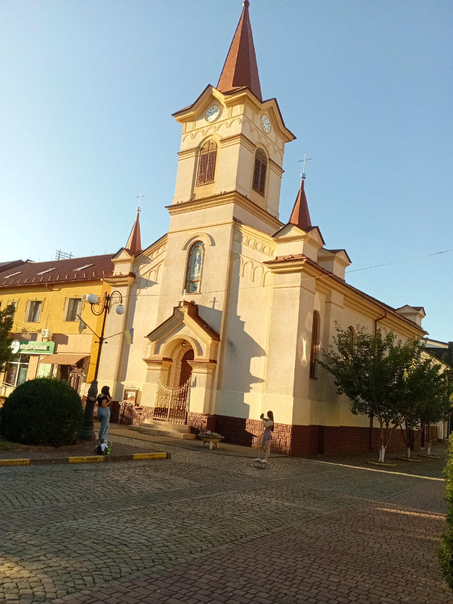 The image captures a religious building, likely a church, in Tiachiv, Ukraine. The structure features a tall, cream-colored tower with a red, conical roof, and smaller red-roofed spires. A clock face is visible on the tower. Adjacent to the church is a yellow building with a brown roof, housing a business with a green sign that reads "ПОДОРОЖНИК". The ground in front of the buildings is paved with cobblestones. Two individuals are present: one stands near a streetlamp and a large green bush, and another is further to the right, appearing to take a photograph. The scene is bathed in the warm light of what seems to be either sunrise or sunset, indicated by the long shadows cast on the ground. A small tree stands to the right of the church.