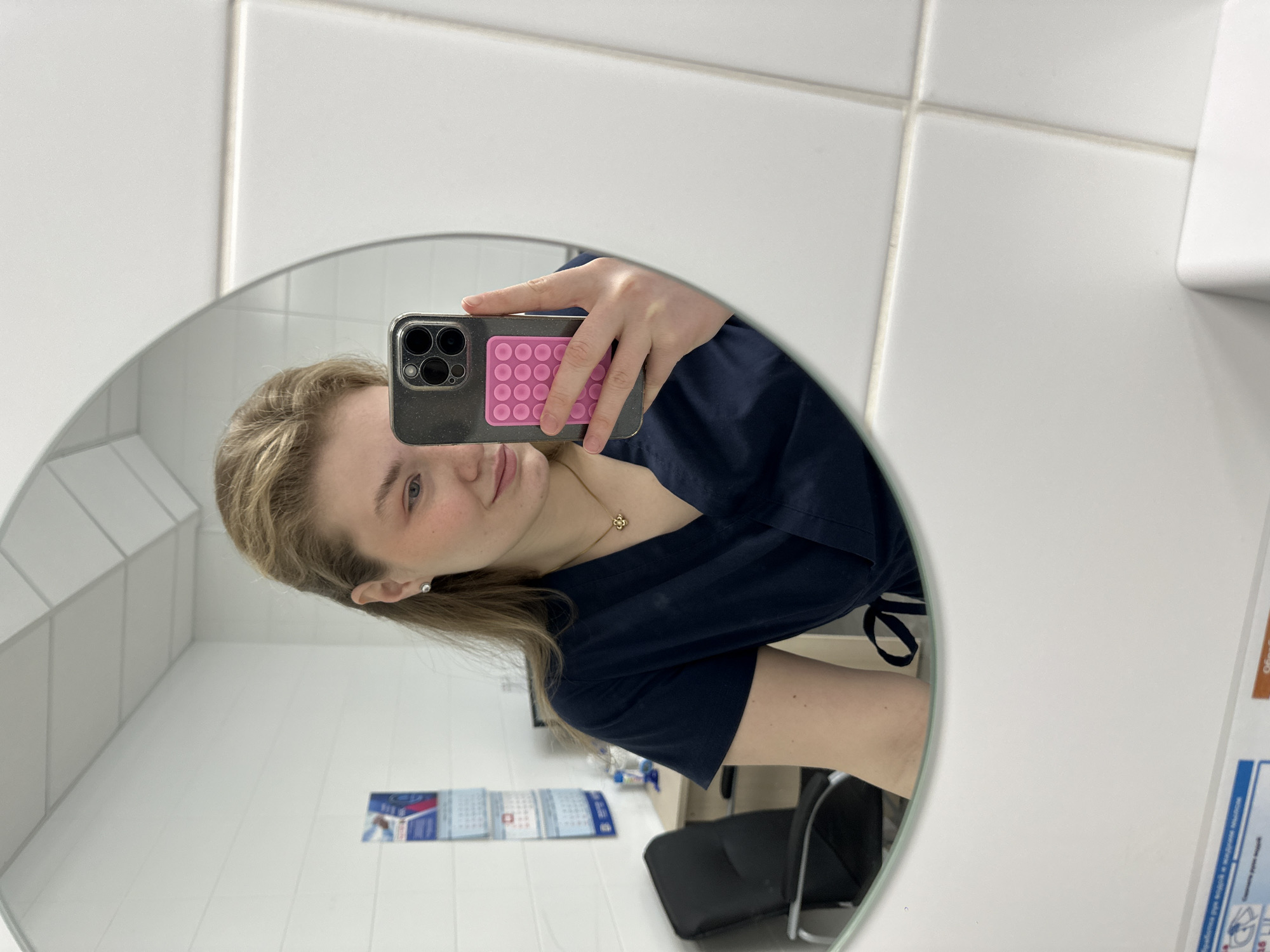 A woman is taking a selfie in a mirror. The mirror is round and framed. She is holding a phone with a gray case and a pink "pop-it" style attachment. She is wearing a dark blue scrub top and a gold necklace with a flower pendant. She has fair skin and blond hair. The background includes white tiled walls, a desk, a black chair, and a calendar in Russian. There is a sign on the wall in Russian. Based on the presence of Russian text and the location of other images like this one, it is likely that this photo was taken in Kazan, Russia.