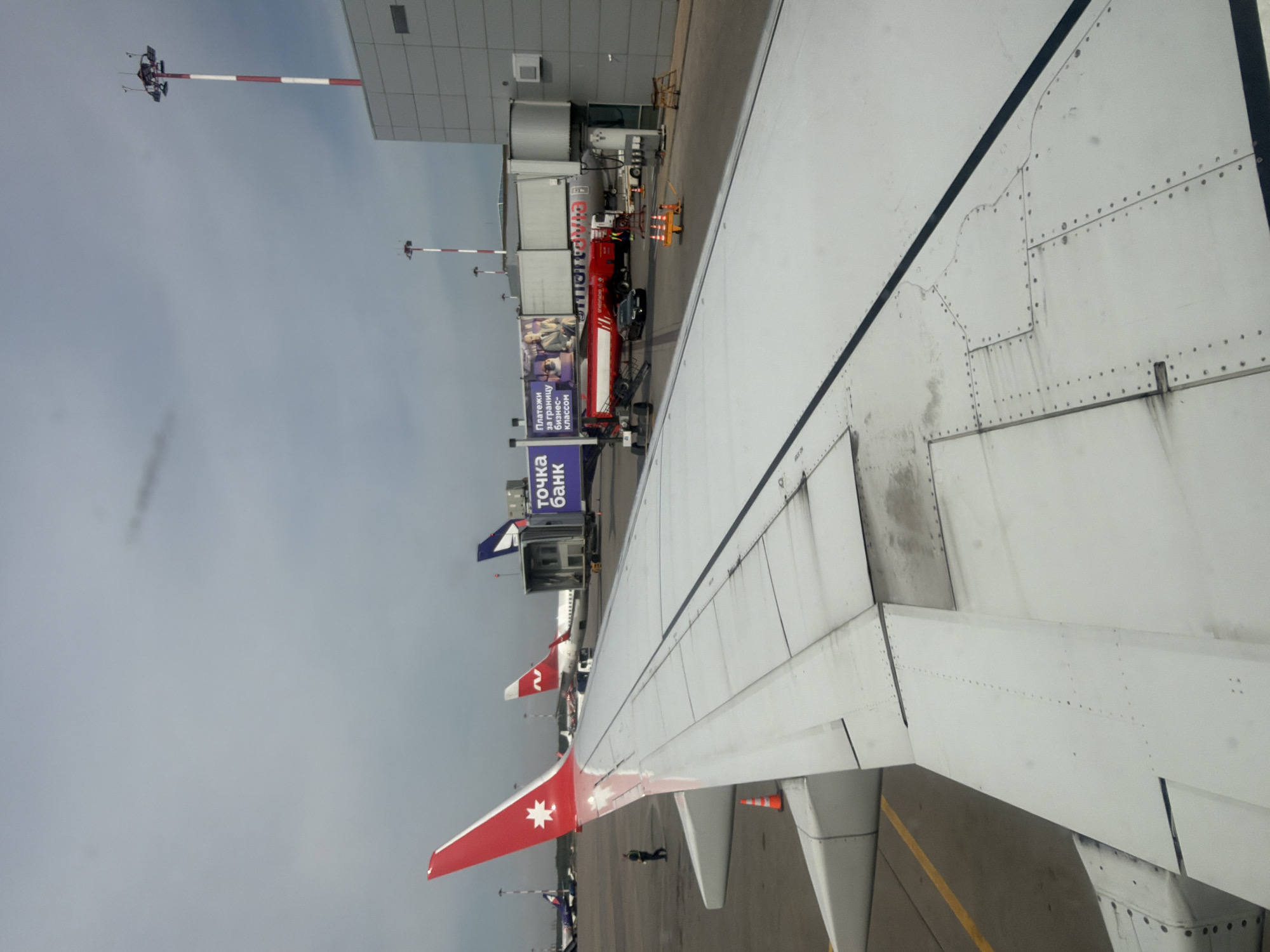 The image shows an airplane wing, presumably taken from inside the aircraft, with a view of an airport scene. The wing is white with visible rivets and a red wingtip featuring a white star. In the background, there are several airplanes parked at terminal gates, some with boarding bridges attached. A red airport service truck is visible next to one of the airplanes, as well as ground personnel. There are also signs with Russian text (including the words "Точка Банк"). The sky is overcast. The image was taken at Pulkovo Airport in Saint Petersburg, Russia.