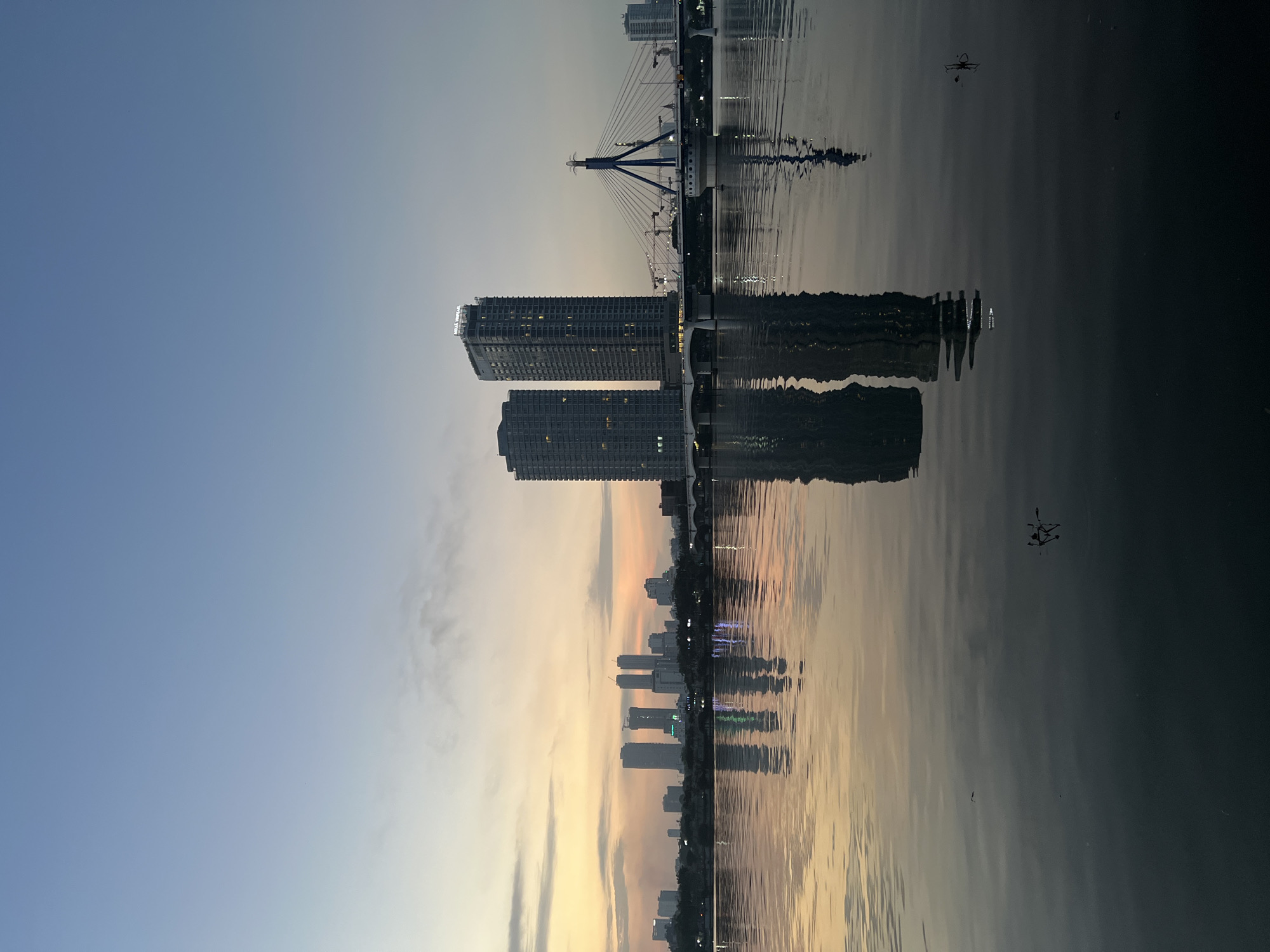 This is an eye-level, full shot of the Han River in Da Nang, Vietnam, during twilight. The sky is a gradient of blue and pale orange, with faint clouds. The city skyline is visible on the horizon, featuring several modern skyscrapers and a cable-stayed bridge. The two most prominent buildings are twin towers, each with many lit windows, reflecting in the still water of the river. The bridge, illuminated by lights, spans the river, with cranes visible behind it, suggesting ongoing construction. The water is dark and calm, mirroring the sky and cityscape, with subtle ripples disturbing the reflections. The overall mood is serene and urban.
