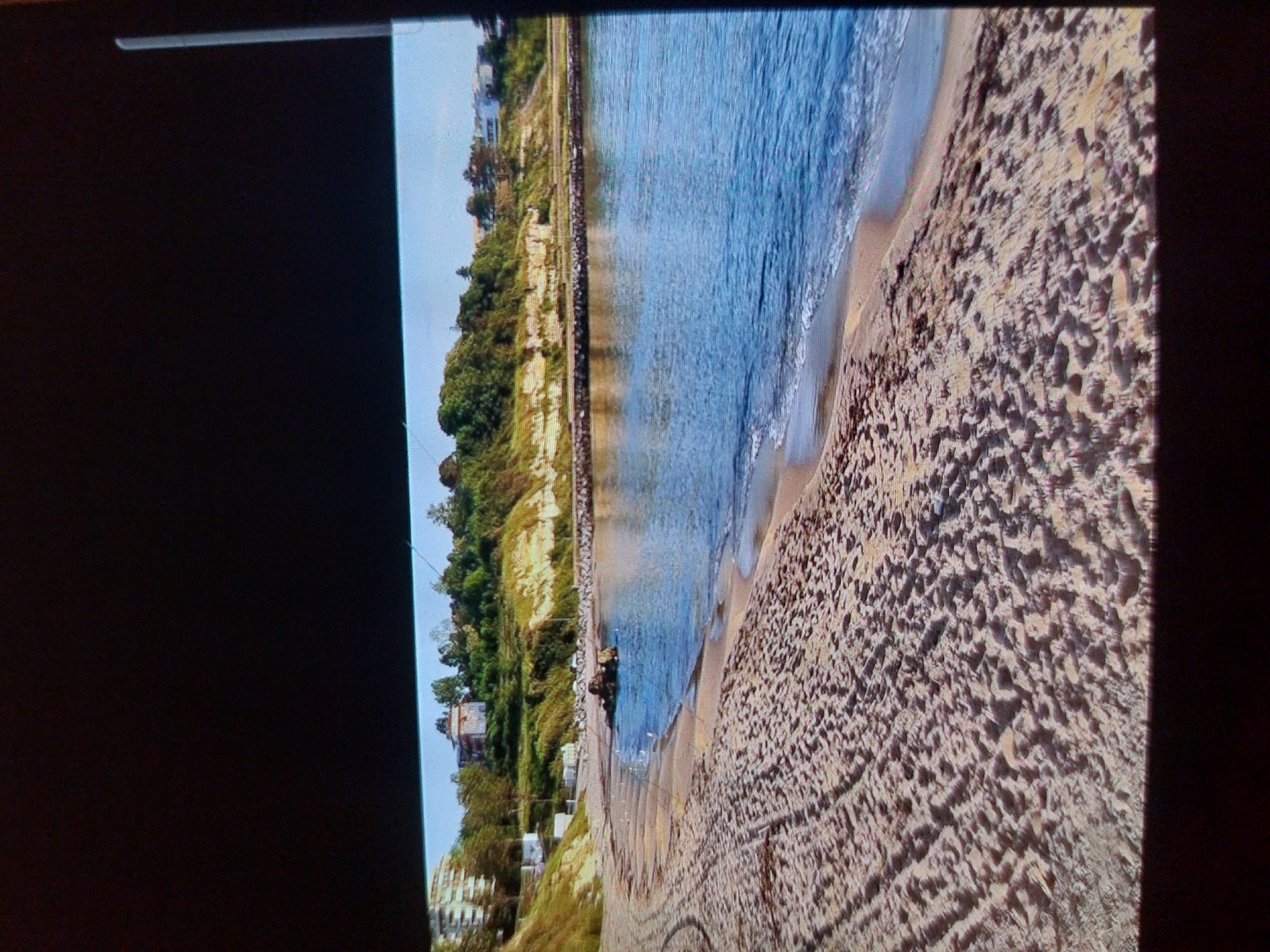 This media file presents a scenic, rotated (90 degrees clockwise) view of a coastal landscape, likely a beach in Tsarevo, Bulgaria.
The image, appearing to be a photograph of a screen, captures a sunny daytime scene with a clear blue sky. In the foreground, a wide sandy beach stretches, displaying intricate textures and ripples, possibly formed by receding waves or wind patterns. No footprints or people are visible on the sand.
Adjacent to the beach is a calm body of blue water, presumably the sea or a large bay. Gentle waves can be seen breaking close to the shore, creating a lighter band of shallow water before deepening to a richer blue. A long, dark, linear structure, possibly a breakwater or a pier, extends parallel to the coastline in the water.
The background features a steep, verdant coastline, characterized by lush green vegetation, including trees and bushes, covering what appears to be cliffs or hills. Nestled among the greenery are several buildings. One notable building, slightly left of center, has a distinctive red roof and appears to be an older structure. Further along the coast to the left, another building with white columns is partially visible.
The scene conveys a peaceful and natural ambiance. No explicit activities or people are discernible. There is no visible text in the image. The black borders on the left and right sides of the image, along with a thin white line at the top left, suggest the picture was taken of a display screen.