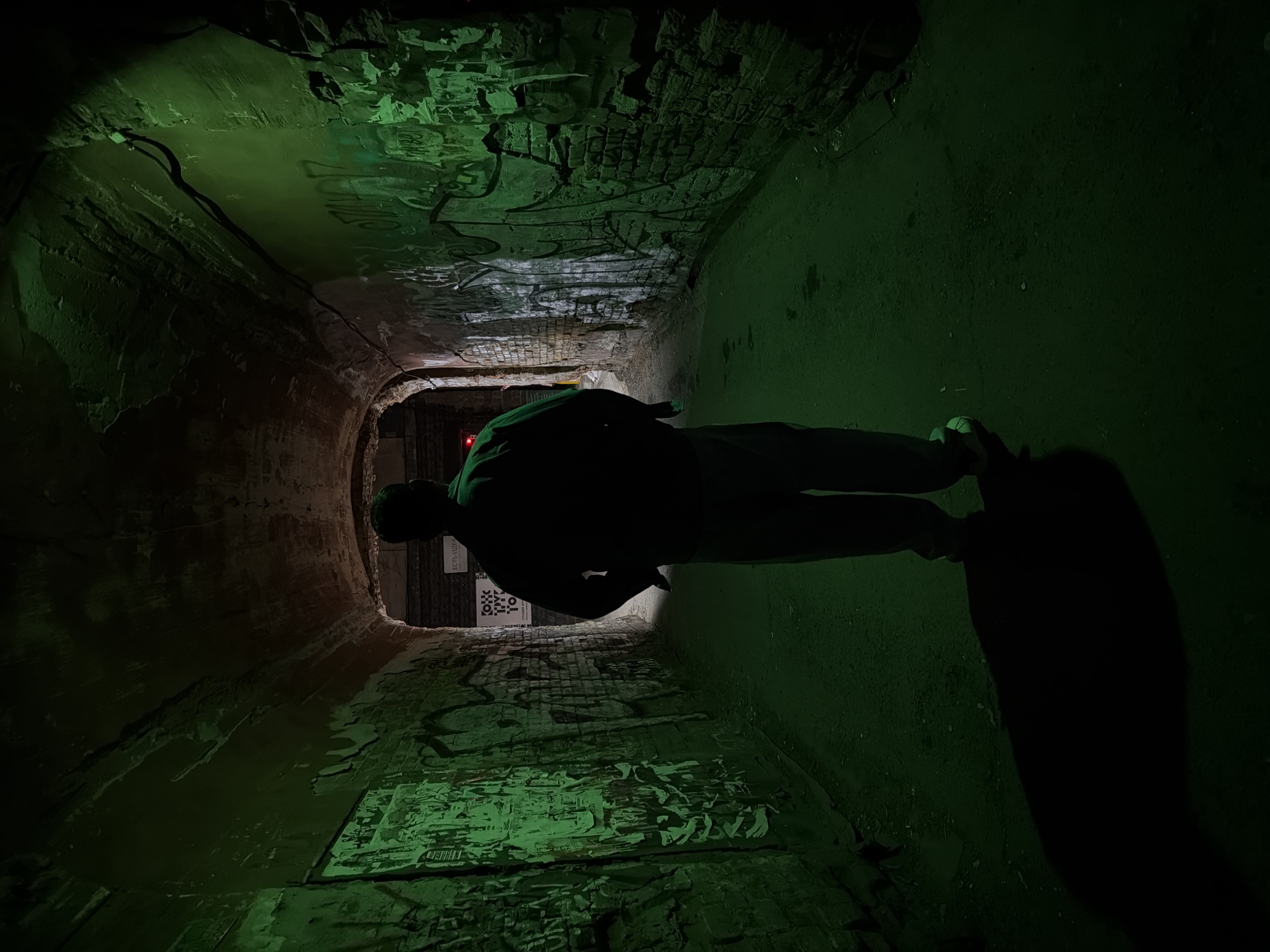 A person stands silhouetted in a dimly lit, tunnel-like passage. The tunnel walls and ceiling display exposed brick and decaying plaster, marked with graffiti and peeling posters. The scene is illuminated by a green-tinted light source, casting long shadows. A red light is visible at the far end of the tunnel. The location is an underpass near the embankment of the Yenisei River in Krasnoyarsk, Russia.
