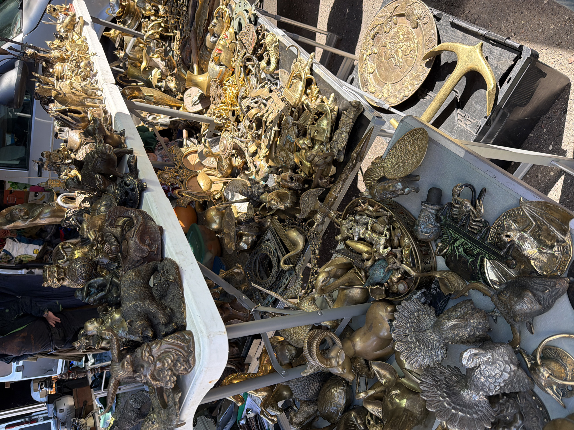 The image displays an outdoor market stall in Riga, Latvia, heavily laden with a vast collection of metallic objects, predominantly brass and bronze figurines and decorative items. The merchandise is distributed across two white folding tables and on the ground level around and beneath them.
On the upper table, an assortment of animal and mythological figures is prominently featured, including a large brass tiger-like creature, a detailed bear figurine, a seated Buddha, a dragon, an eagle with spread wings, and smaller statues of oxen and turtles. The table's surface continues to the right with numerous smaller brass animal figures, such as elephants, deer, and various decorative items.
Below the tables and on the ground, additional metallic objects are arranged. These include larger figurines of rams, turkeys, swans, and ornate metallic plates, one large circular plate showcasing embossed mythological or classical scenes. A golden anchor rests on a black plastic storage crate on the right side of the image. The ground also holds decorative, empty metallic frames and a large quantity of tangled brass chains, handles, and miscellaneous hardware. A person, likely a vendor, is partially visible in the upper left, standing behind the table, wearing a dark jacket and a t-shirt with a graphic design. Bright sunlight illuminates the scene, creating distinct shadows on the ground, which appears to be reddish-brown packed earth or gravel. A white commercial van with its rear doors open is parked directly behind the stall, suggesting its use for transporting the goods.
