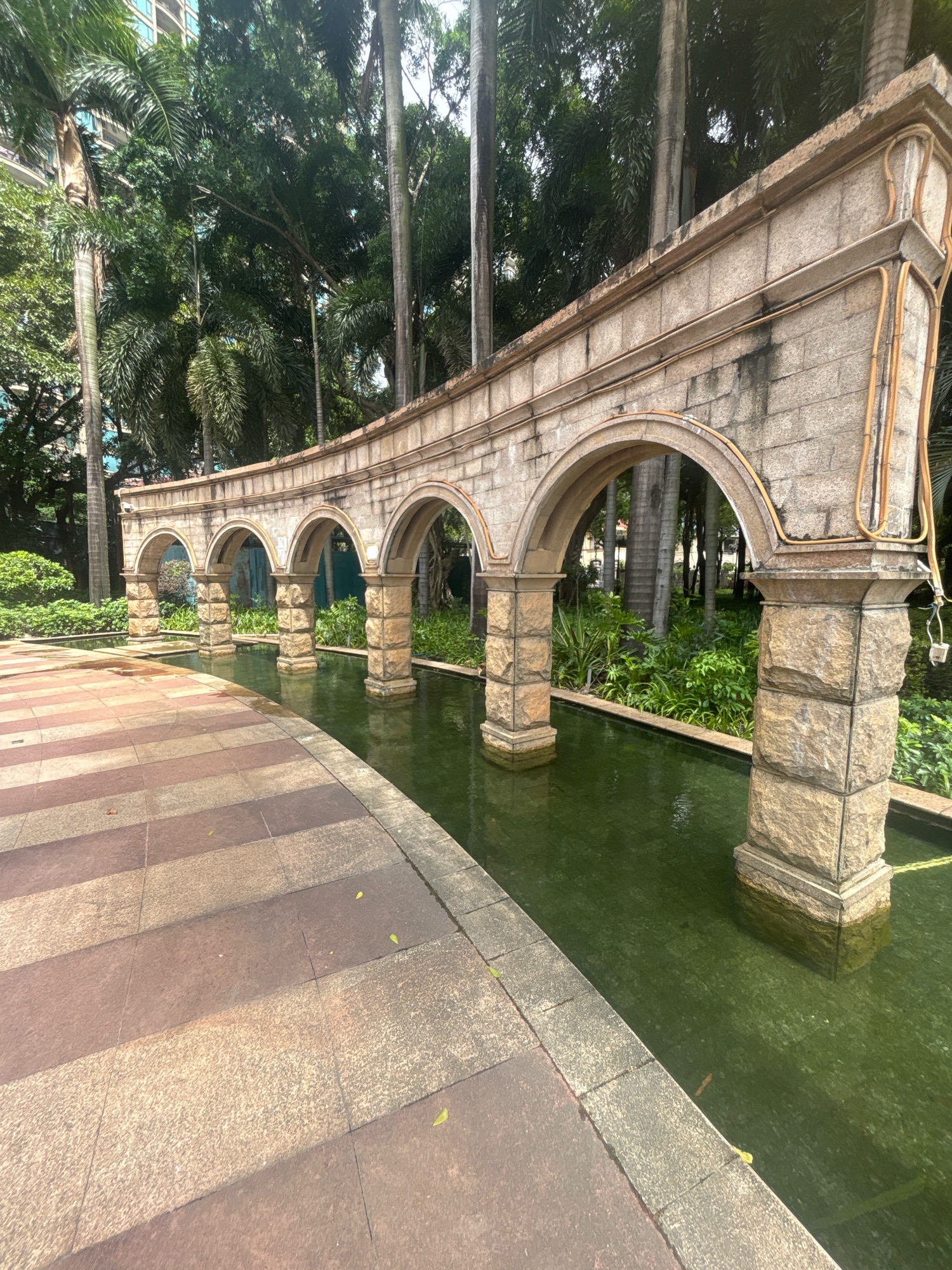 The image depicts an outdoor scene featuring a stone arcade with multiple arches bordering a narrow, shallow pool of green water. The arcade is constructed from light-colored stone blocks and has a decorative, curved top. Several palm trees and lush green foliage are visible behind the arcade. In the foreground, a pathway made of rectangular stone tiles in varying shades of brown and beige runs alongside the water feature. The lighting suggests it is daytime, with sunlight casting shadows on the pathway. The environment appears to be a park or a garden within a city setting, given the presence of tall trees and hints of a modern building in the upper left corner. The scene is devoid of any people or animals. This is an image of the "Archway of the Hundred Arches" located in the Nanhai District of Foshan City, China.