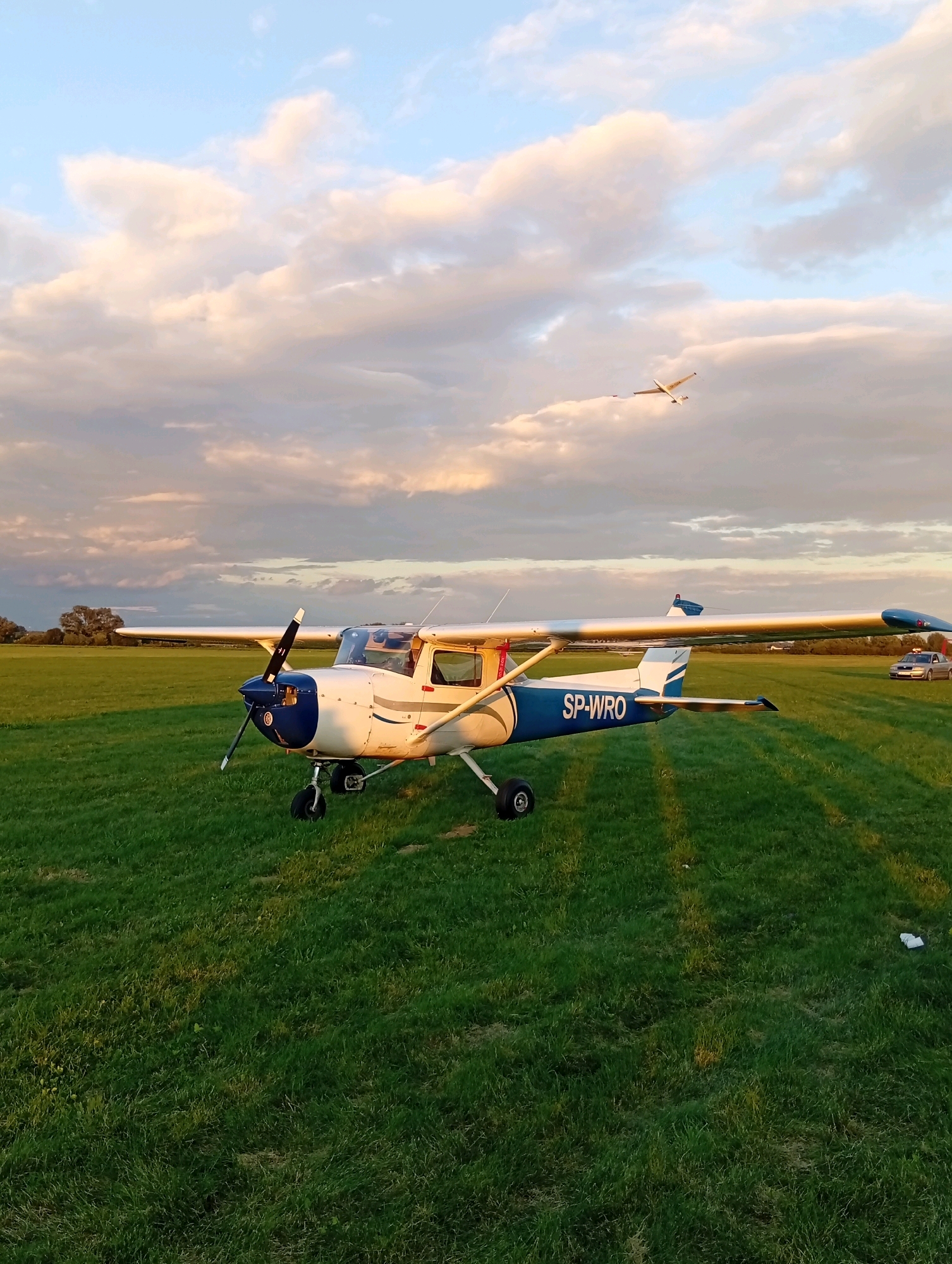 A small white and blue propeller airplane with the registration "SP-WRO" is parked on a grassy airfield. The plane has its propeller visible. In the background, there is a field of green grass stretching towards a line of trees. The sky is a mix of blue and fluffy white and pink clouds, indicating either sunrise or sunset. Another small, distant aircraft is visible flying in the upper right portion of the sky. A silver car is parked in the distance on the right side of the airfield. The image appears to have been taken in Szewce, Poland.
