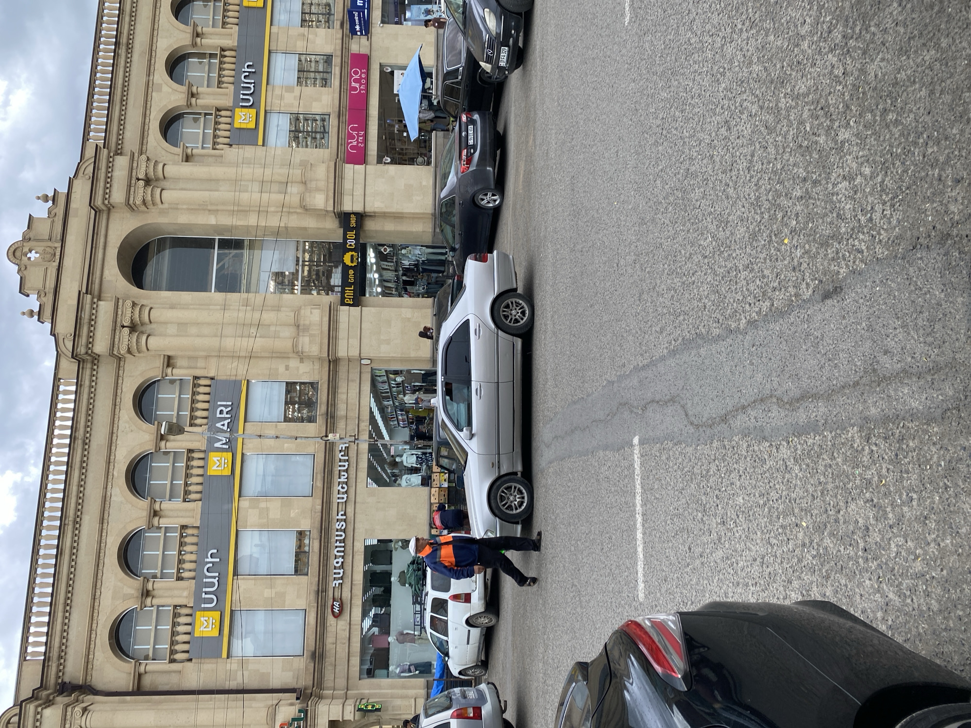 The image captures a street scene in Gyumri, Armenia, in front of a building with multiple storefronts. The building features a beige, classical architectural style with arched windows and decorative columns. Prominent signs indicate businesses such as "MARI" and "Uno shoes." A man in a blue and orange jacket and black pants is walking across the street. Several cars are present on the asphalt road, including a silver sedan, a white SUV, a dark grey sedan, and a black car partially visible in the foreground. The sky is overcast. The environment suggests a typical urban setting.