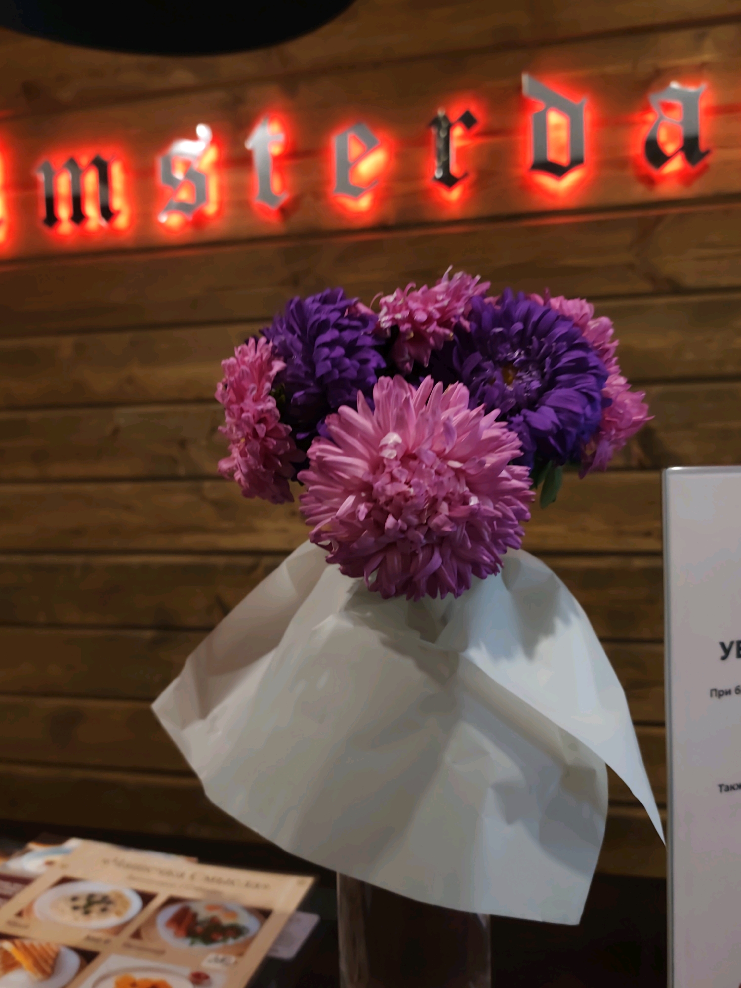 The image shows a bouquet of pink and purple asters in a white wrapper, placed in front of a wooden wall with a neon sign that spells "amsterda". The sign appears to be red and has a gothic font. To the right of the bouquet, a white sign with Cyrillic text is partially visible. At the bottom of the image, a blurred menu with images of food is visible. The scene is likely in a restaurant or cafe in Kazan, Russia, given the Cyrillic text and the style of the signage, which is reminiscent of places that may exist in the city. There are no people or animals visible in the image. The lighting suggests an indoor setting.
