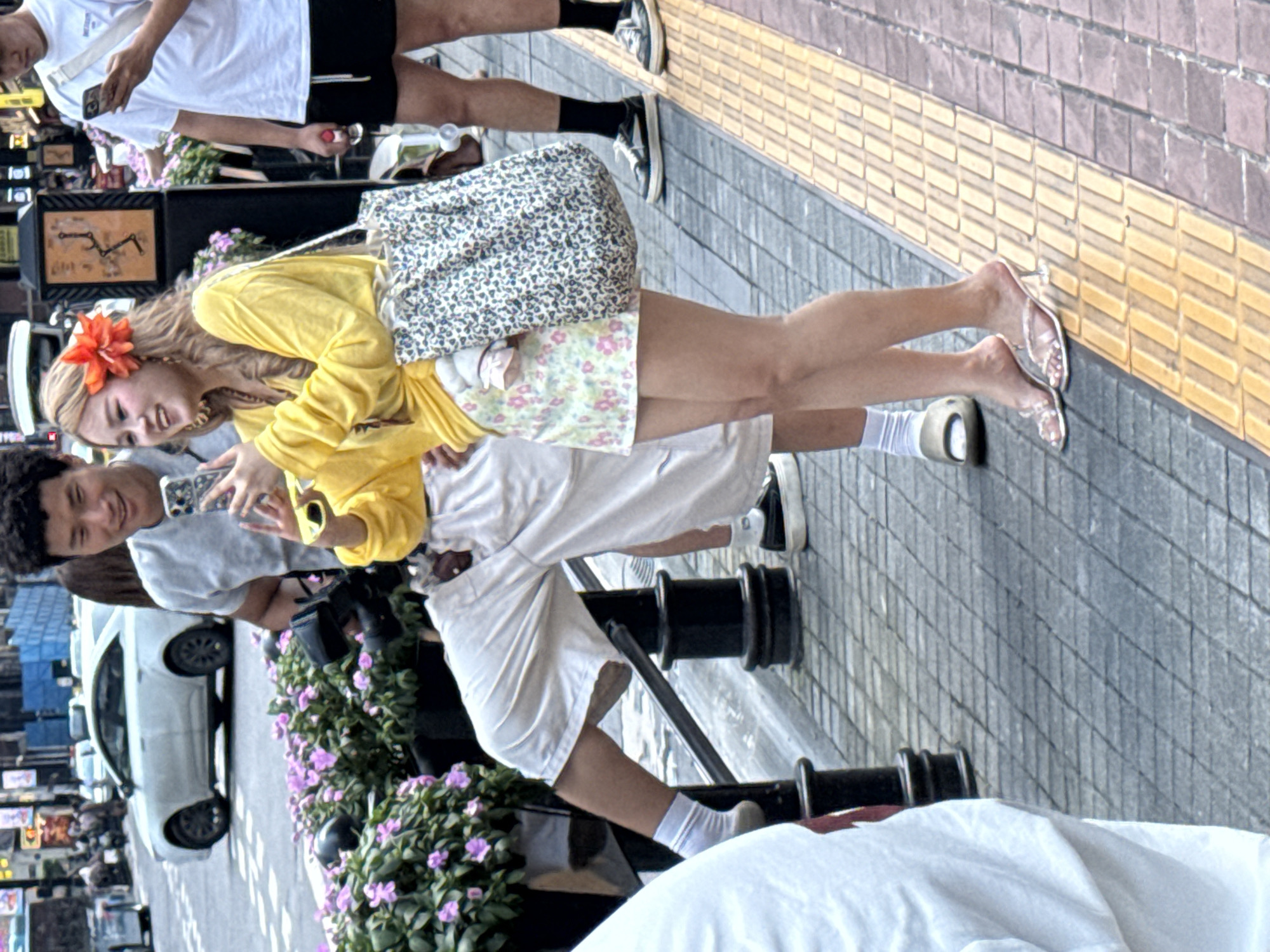 In Yuzhong District, China, two people stand on a sidewalk next to a railing. The person on the left is wearing a white t-shirt and white shorts, with white socks and black sandals. They are holding a phone in their right hand. The person on the right is wearing a yellow long-sleeved shirt, a floral print skirt, and clear heeled sandals. They have a red flower in their blonde hair and are holding a phone in their right hand, which is pointed towards the person on the left. A patterned tote bag hangs from their left shoulder. Behind them, a white car is visible on a street, along with a white and black railing.