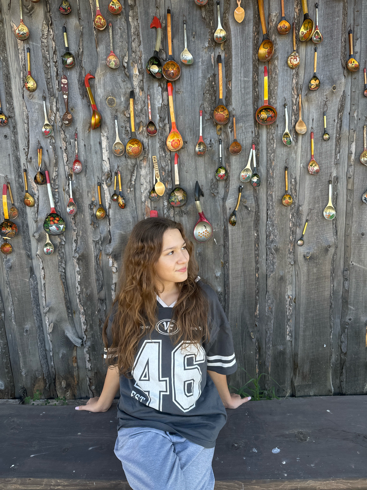 In the city of Kargat, Russia, a young woman with long, wavy brown hair sits on a dark wooden bench. She is wearing a charcoal grey t-shirt with the number "46" emblazoned in white, along with a white-striped collar and sleeve cuffs. Her light grey sweatpants are partially visible. The woman is looking to her right, her gaze directed off-camera. Behind her, a weathered wooden fence serves as a backdrop, adorned with numerous brightly painted wooden spoons. These spoons, showcasing a variety of traditional Russian folk art designs, are hung in vertical and diagonal rows across the fence. The lighting suggests it is daytime, with natural light illuminating the scene.