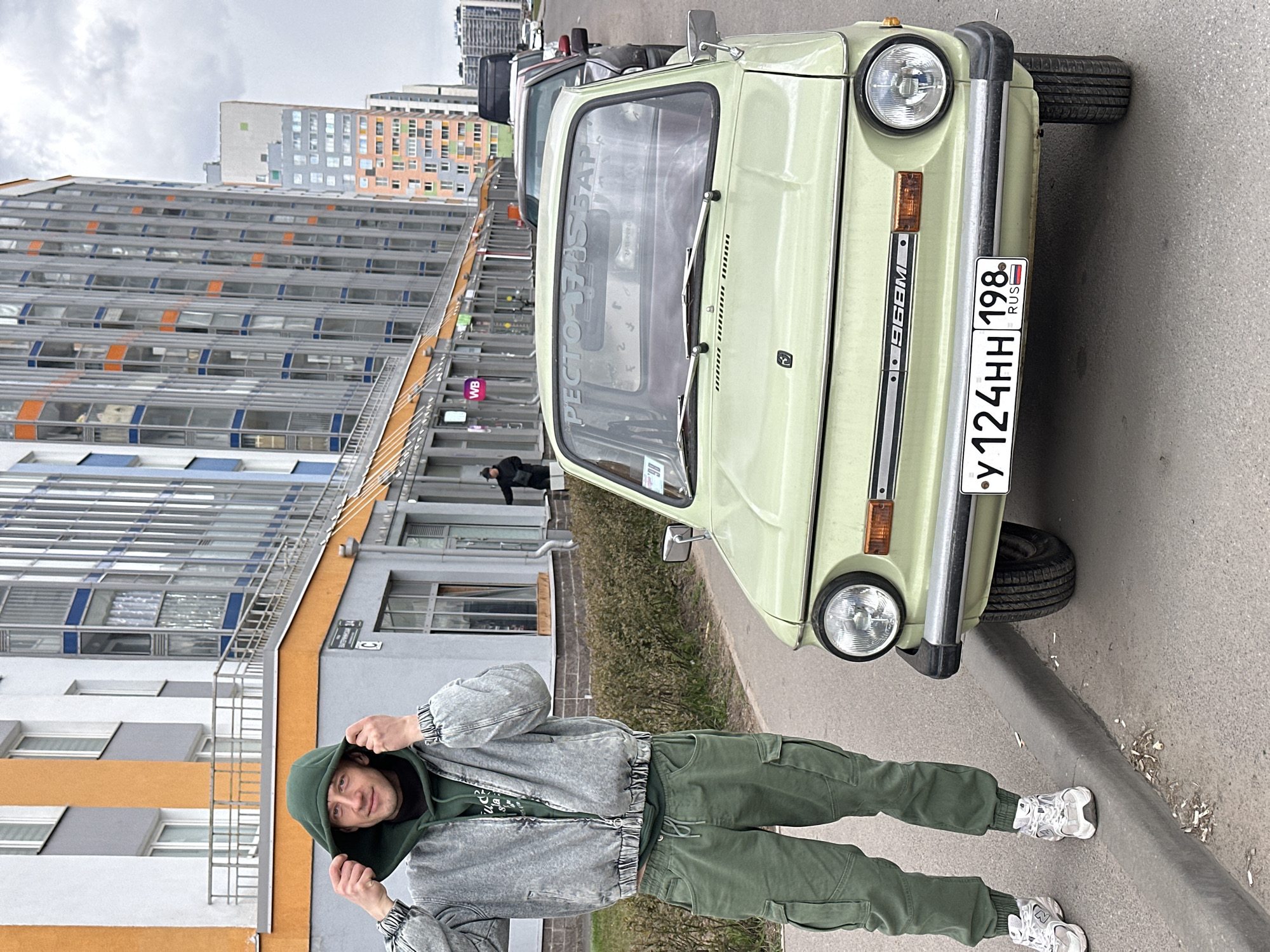 This media file depicts a young man posing next to a vintage light-green car on a street in Murino, Russia, under an overcast sky.
The man, positioned in the foreground to the left, wears a dark green hooded sweatshirt, a light gray distressed denim jacket, and olive green cargo pants, completed with white sneakers. He is looking towards the viewer with a slight smile, holding the hood of his sweatshirt with both hands.
To the man's right, a classic light-green Soviet-era car, likely a ZAZ-968M (indicated by "968M" on its side trim), is prominently featured. The car has distinctive round headlights, a chrome bumper with black accents, and a black front grille. Its Russian license plate, "У 124 НН 198 RUS," identifies it as registered in the Saint Petersburg region (198). Reflected in the car's windshield, partially obscured, is text that appears to read "РЕСТОРАН ТЬЮБАР" (Restaurant Tube Bar).
In the background, several modern, multi-story apartment buildings with varied facades of orange, blue, gray, and white panels line the street, characteristic of contemporary residential developments in Russia. Sparse green bushes are visible between the street and the buildings. A purple square sign with "WB" (likely Wildberries, a Russian e-commerce company) is faintly visible on one of the buildings. Other parked vehicles can be seen further down the street. The overall lighting is soft and diffused, characteristic of an overcast day. The setting suggests an urban residential area.