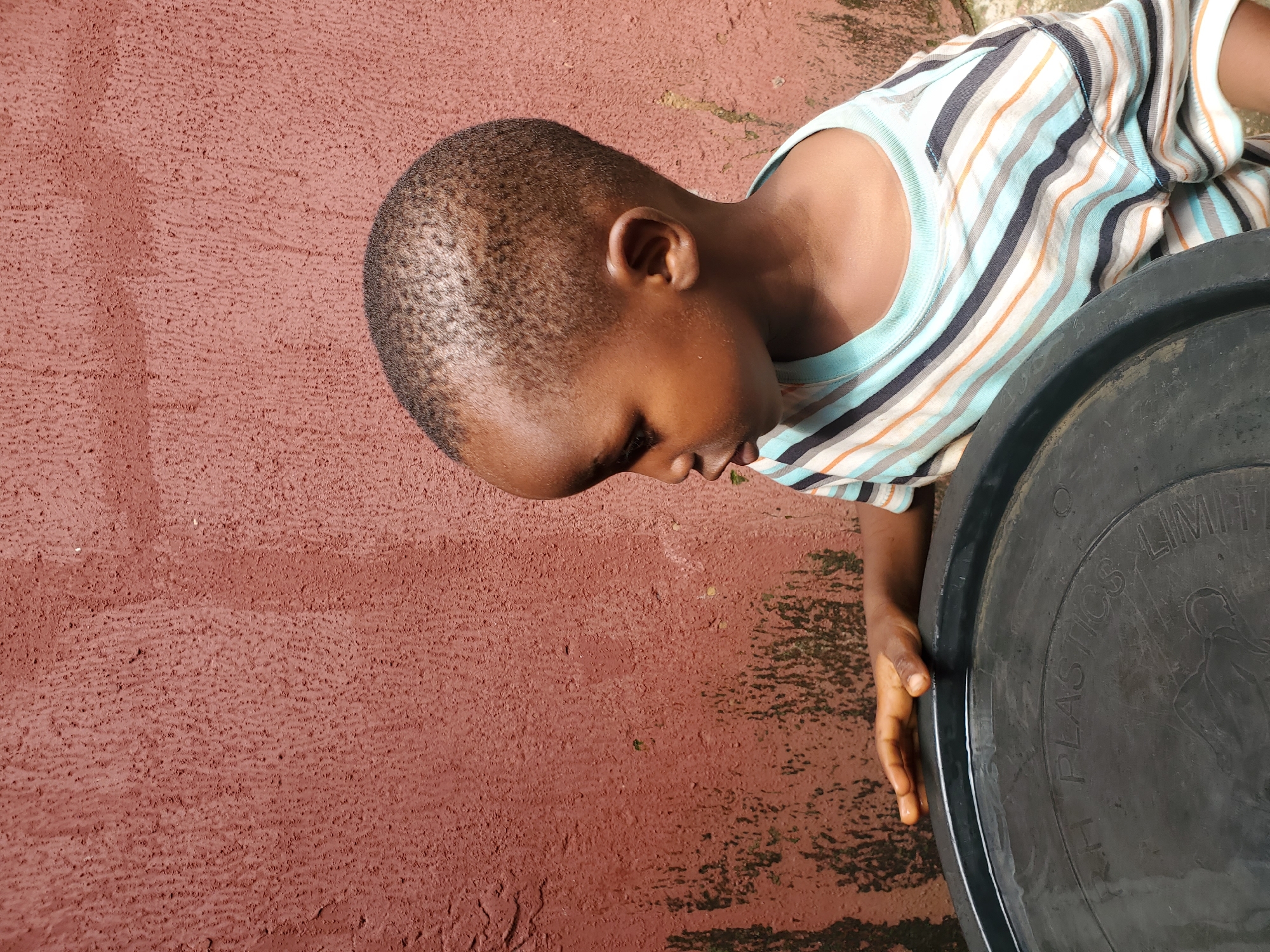 In this medium shot, a young Black child with short hair is shown from the side, looking downwards. The child is wearing a horizontally striped t-shirt with thin bands of white, light blue, dark gray, and orange. They are positioned next to a large, black plastic container, the rim of which takes up the right side of the frame. The child's left hand rests on the edge of the container.
The setting appears to be outdoors, with a reddish-brown textured surface occupying the left and background areas. This surface has some darker, possibly damp patches, suggesting it might be wet or weathered. The overall lighting suggests it is daytime, possibly late morning or afternoon, given the moderate shadows. The scene conveys a sense of calm or quiet contemplation from the child.
Visible text on the black container reads "RH PLASTICS LIMIT" and what appears to be "ED". The context of Jos, Nigeria is provided for this image.