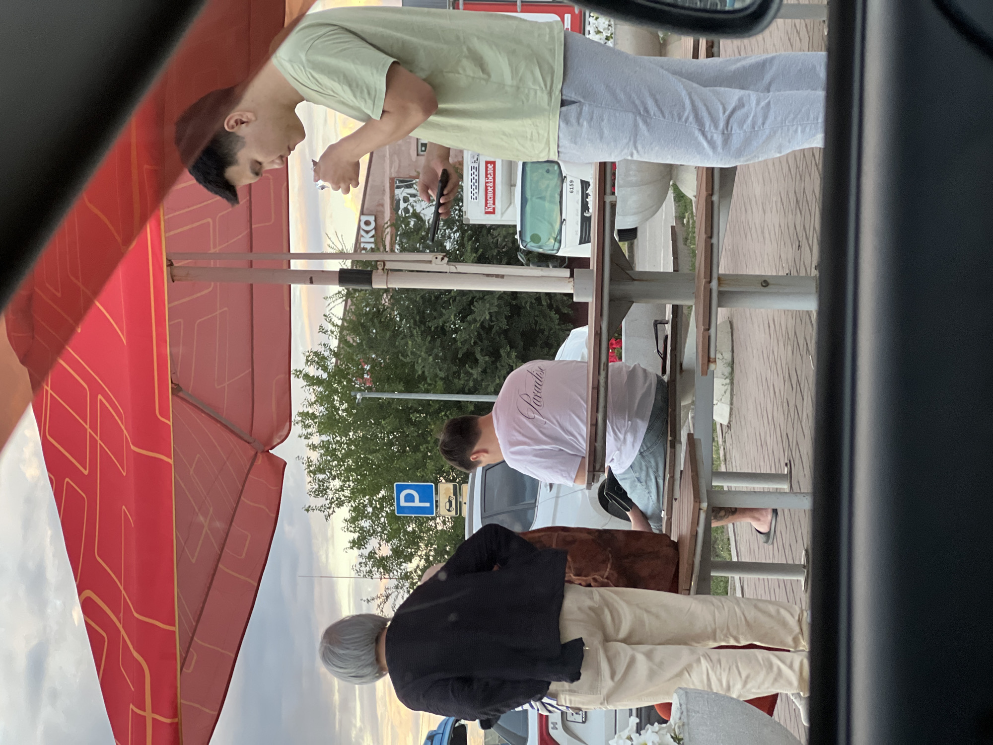 The image, taken in Zimovniki, Russia, shows three individuals in an outdoor setting, likely a cafe or rest area with outdoor seating. The time of day appears to be late afternoon or early evening, with a soft, warm light suggesting sunset.
In the foreground, a person with light gray hair and a black jacket is facing away from the camera, wearing light-colored pants. To their right, a person wearing a white t-shirt with the word "Paradise" printed on the back and light blue jeans is seated. They are looking down, and their legs are crossed. The third person, in the background, is wearing a light green t-shirt and light gray pants. They are also looking down and holding a phone in one hand and what appears to be a cigarette in the other.
The scene is set with tables and chairs, typical of an outdoor dining or resting area. A red awning with a yellow geometric pattern is visible on the left. In the background, there are trees, a white truck with Russian text that reads "Красное & Белое" (Red & White), and a parking sign with a blue "P". The ground is paved with bricks. The overall mood seems relaxed and casual.
