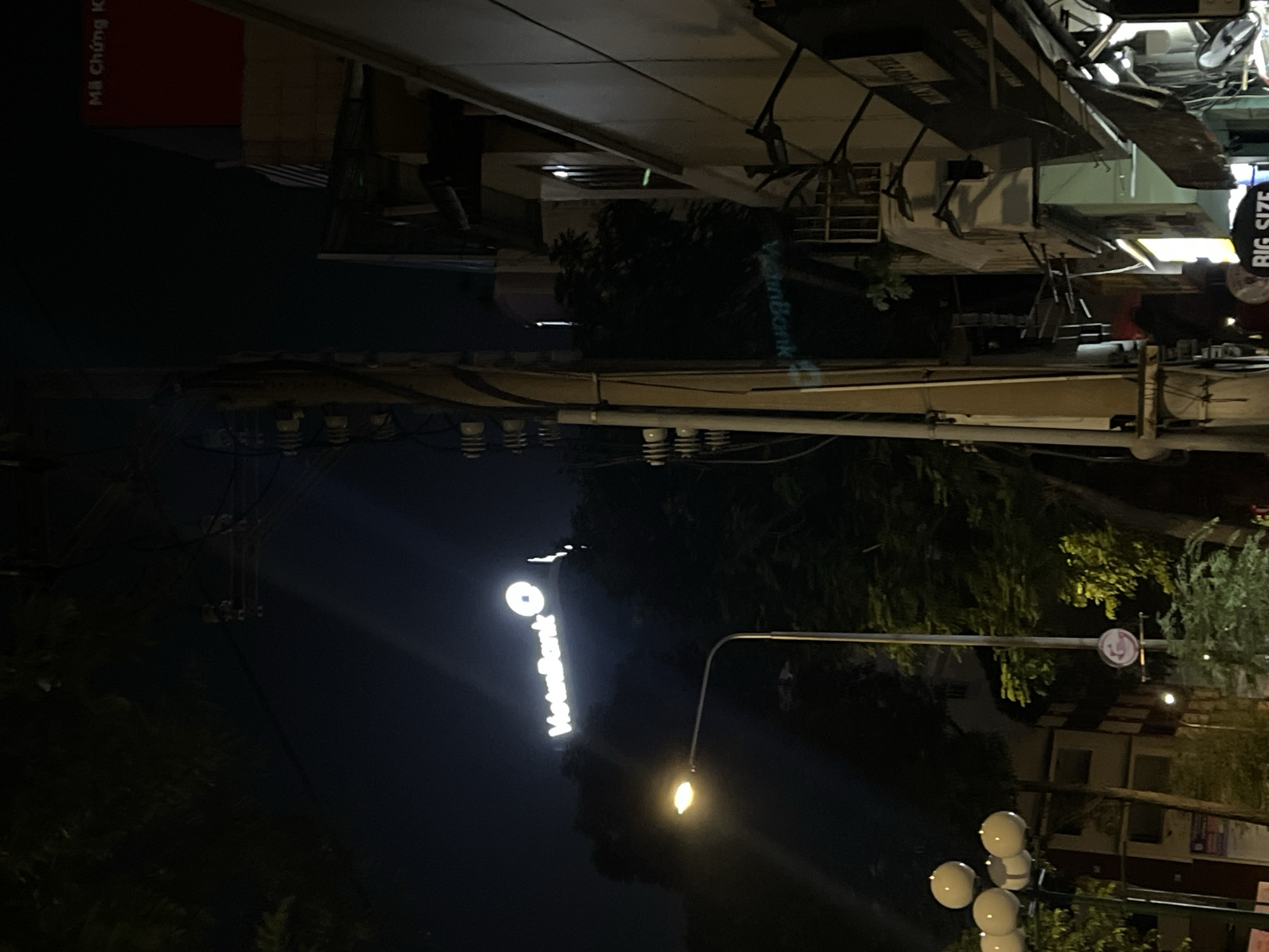The image shows a night scene in Thủ Đức, Vietnam. A lit "NodataBank" sign is visible vertically on the left side of the frame. Streetlights illuminate a street lined with trees and buildings. Overhead power lines crisscross the sky. A building with a "BIG SIZE" sign is located on the upper right. There are other street-level signs on other buildings as well.