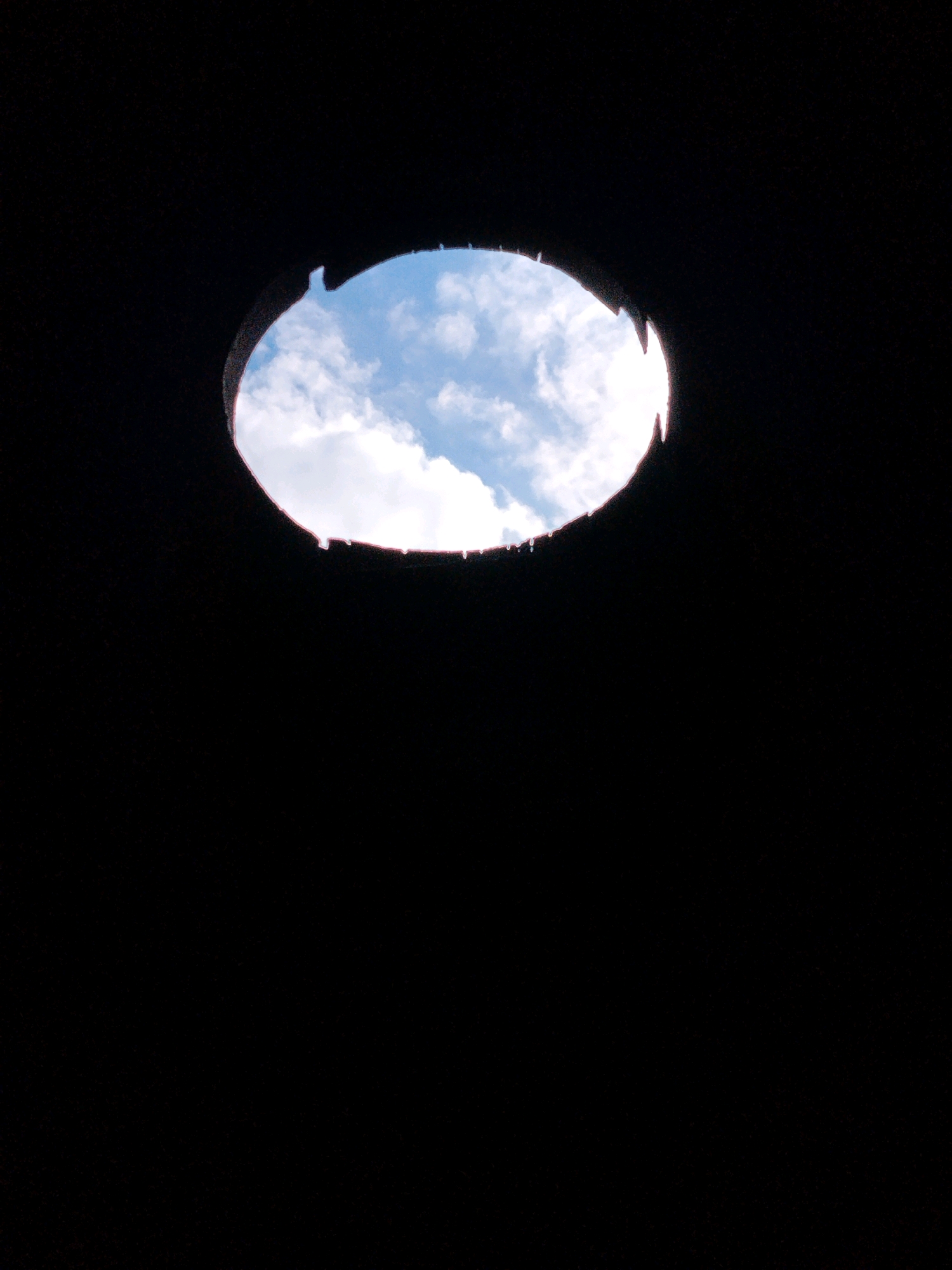 The image shows a circular opening in a dark ceiling, revealing a bright blue sky with white clouds. The edges of the opening are uneven and appear to be made of a dark material, possibly metal or concrete, with some jagged protrusions. The opening is centrally located in the upper portion of the frame, against a background of deep black. The overall impression is that of looking upwards through a skylight or an aperture in a roof. There are no people, animals, or other discernible objects visible within the frame. The scene is devoid of any specific environmental context that would suggest its location in the city of Hoholove, Ukraine, or any other specific urban or natural setting. The lighting suggests it is daytime.
