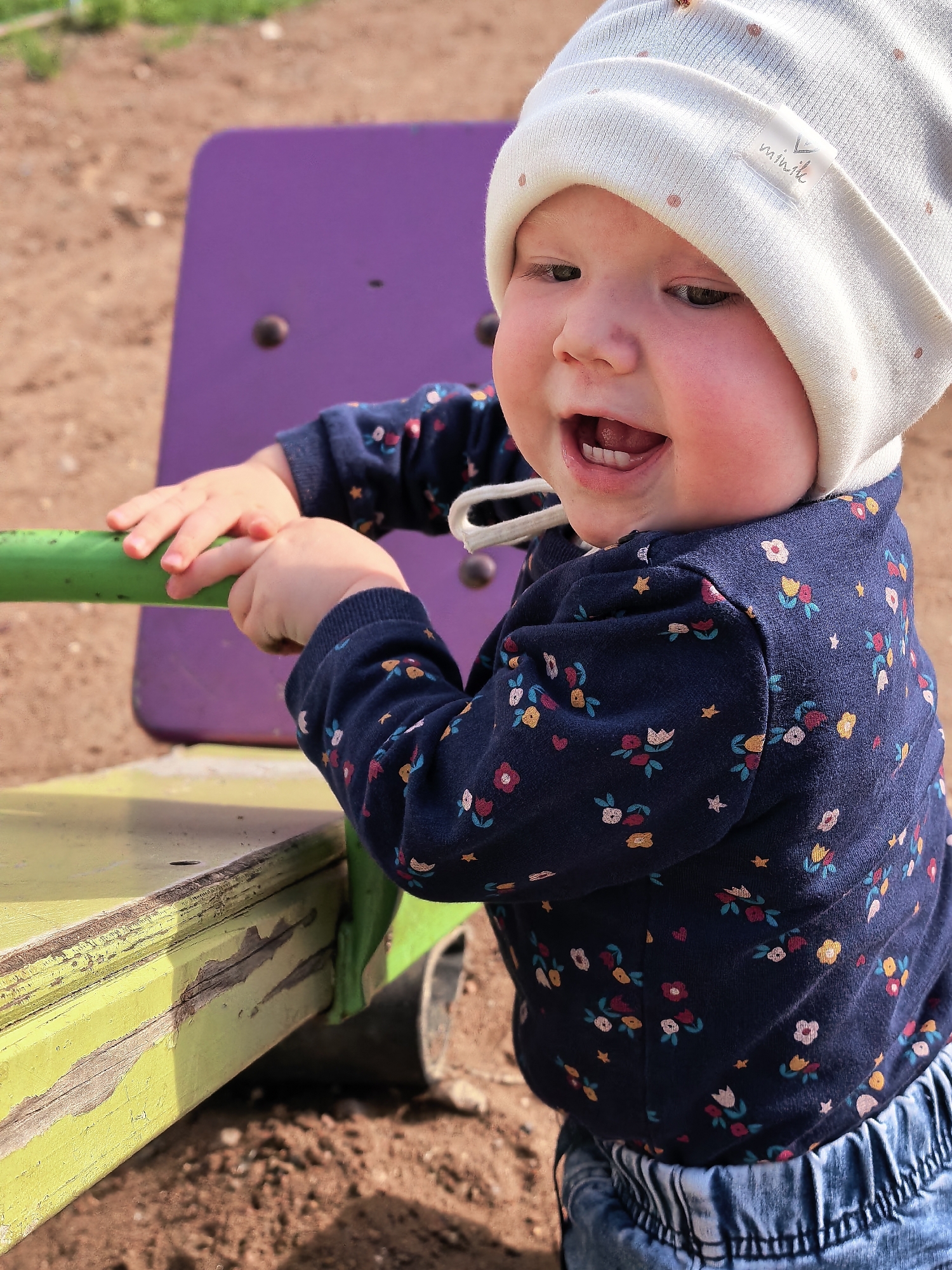 An outdoor close-up depicts an infant or toddler engaging with playground equipment. The child, with fair skin and rosy cheeks, is positioned in the foreground, looking slightly to the viewer's left with an open-mouthed expression suggesting laughter or vocalization. The child wears a light cream-colored knitted beanie featuring small, light brown polka dots; a white fabric label with the stylized text "minile" is affixed to the right side of the beanie. A white drawstring is visible hanging from the beanie. The child's torso and arms are clad in a dark blue long-sleeved shirt patterned with small, multi-colored flowers and light-colored stars. The lower portion of the child's body shows light blue denim pants with an elasticized waistband. Both of the child's hands grip a horizontal, cylindrical green bar, which is part of a piece of playground apparatus. Beneath this green bar, a worn, rectangular structure, painted a chipped lime green, is visible, revealing a darker underlying material. In the mid-ground, a flat, upright structure painted solid purple, featuring two dark circular fasteners or caps, stands behind the child. The ground surface is composed of reddish-brown dirt or sand. The scene is brightly lit by natural sunlight. The image contains no visual elements to confirm the specified location in Sterlitamak, Russia.