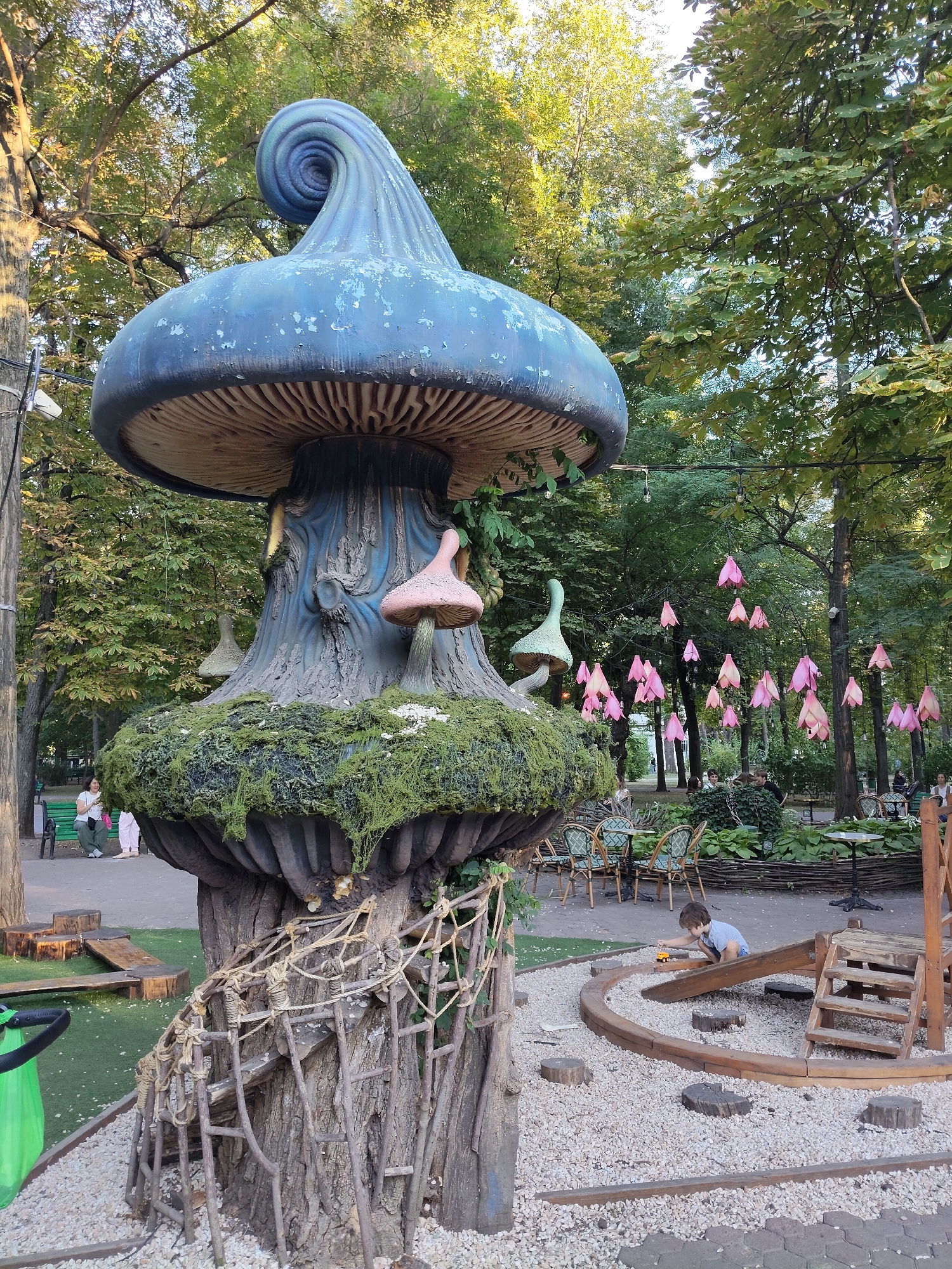 The image depicts a whimsical outdoor scene, likely a park or garden, with a prominent, oversized mushroom sculpture as the central focus. The sculpture, painted in shades of blue and grey with a swirled top, is adorned with smaller, pink mushroom-like elements. The base of the mushroom sculpture is covered in green moss and has a rustic, tree-trunk-like appearance with rope and wooden ladder accents.
In the foreground, a young child, wearing a blue shirt and shorts, is playing with a toy car on a wooden ramp and track constructed with small tree stumps and gravel. A wooden playground structure with a slide and stairs is visible to the right of the child. The ground is covered with a mix of gravel and green artificial turf.
Behind the mushroom sculpture, the scene opens up to a wooded area with trees and foliage. String lights with pink, bell-shaped decorations are suspended between the trees, creating a festive atmosphere. Several tables and chairs, seemingly for outdoor dining, are arranged in the background, with a few people seated or walking around. A woman is visible sitting on a park bench to the left of the frame. The overall environment suggests a public space designed for recreation and enjoyment, likely during the daytime, with dappled sunlight filtering through the trees.
The provided context indicates that this scene is located in Chișinău, Moldova. Based on the visual elements, this appears to be an area within a park or a themed attraction designed to evoke a fantasy or fairytale aesthetic.