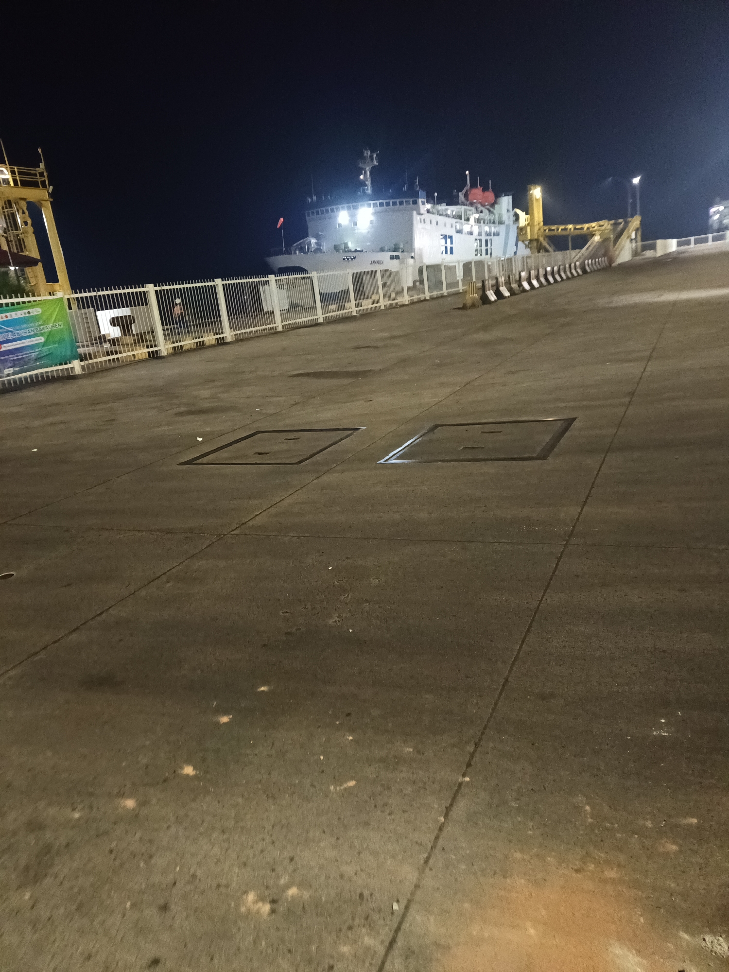 The image shows a concrete surface in what appears to be a port or ferry terminal area at night, likely in Bakauheni, Indonesia. A large white ferry, labeled "AMARISSA," is docked at a ramp, illuminated by lights. A yellow loading structure extends from the ferry to the dock. A white metal fence separates the dock area from the concrete surface in the foreground. There are two square manhole covers visible on the concrete surface. Several scattered white papers or debris are present on the ground. The sky is dark.